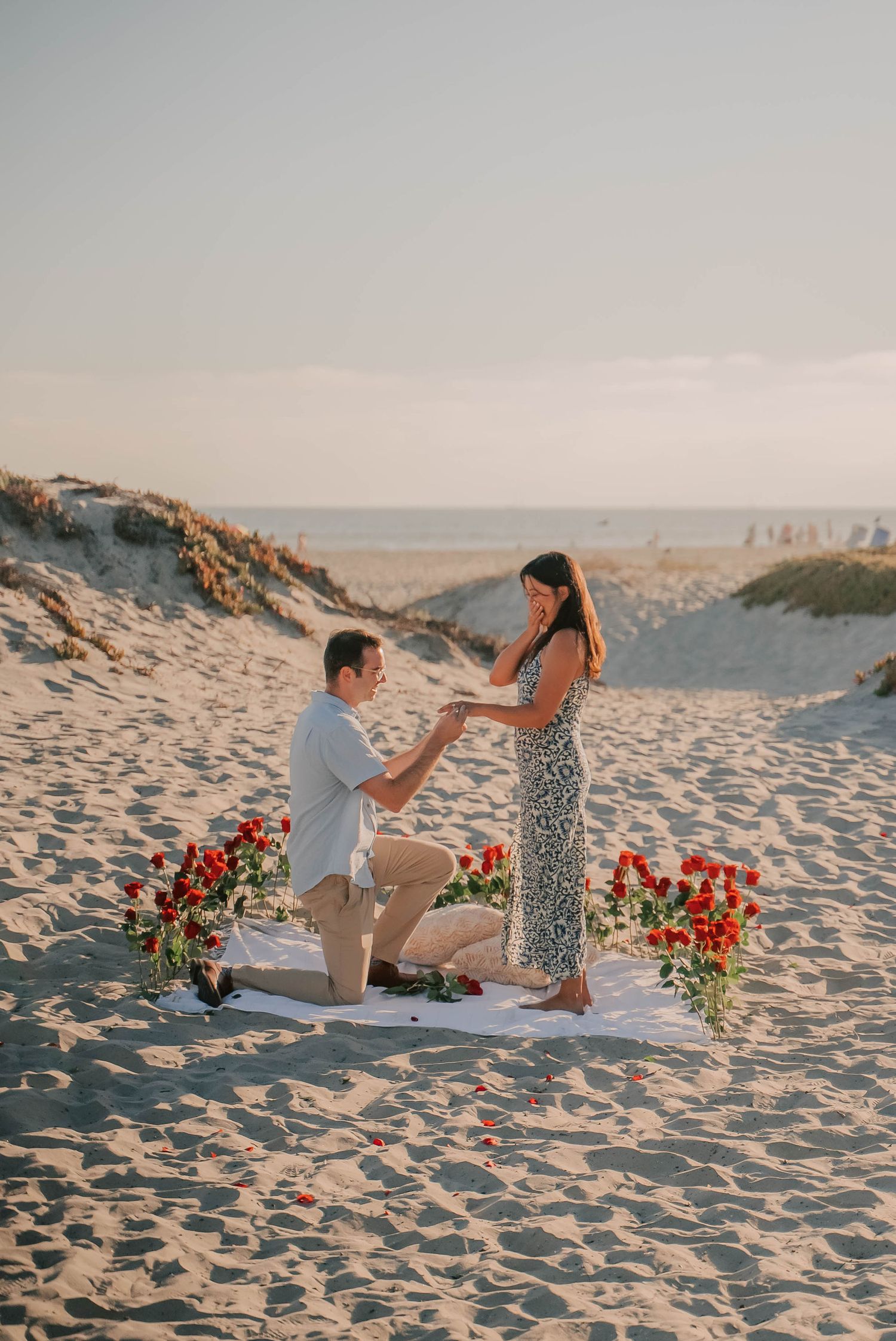 Man proposing on one knee to woman on sandy beach at sunset, surrounded by red roses on white blanket.