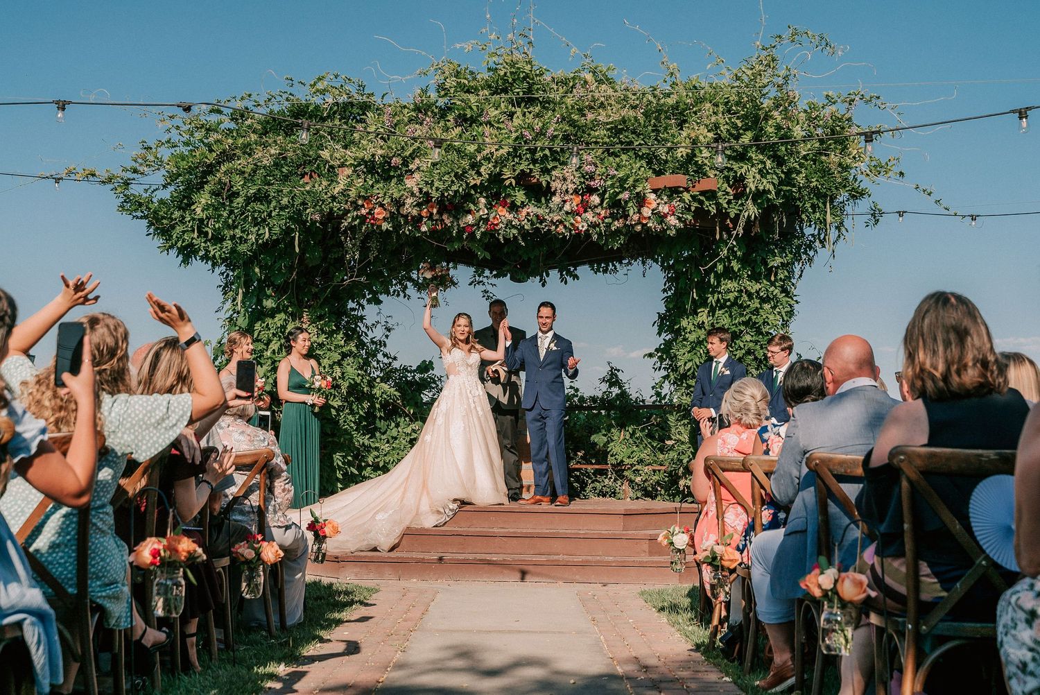 Bride and groom celebrate at outdoor wedding ceremony under floral arch, guests cheering along aisle on sunny day.