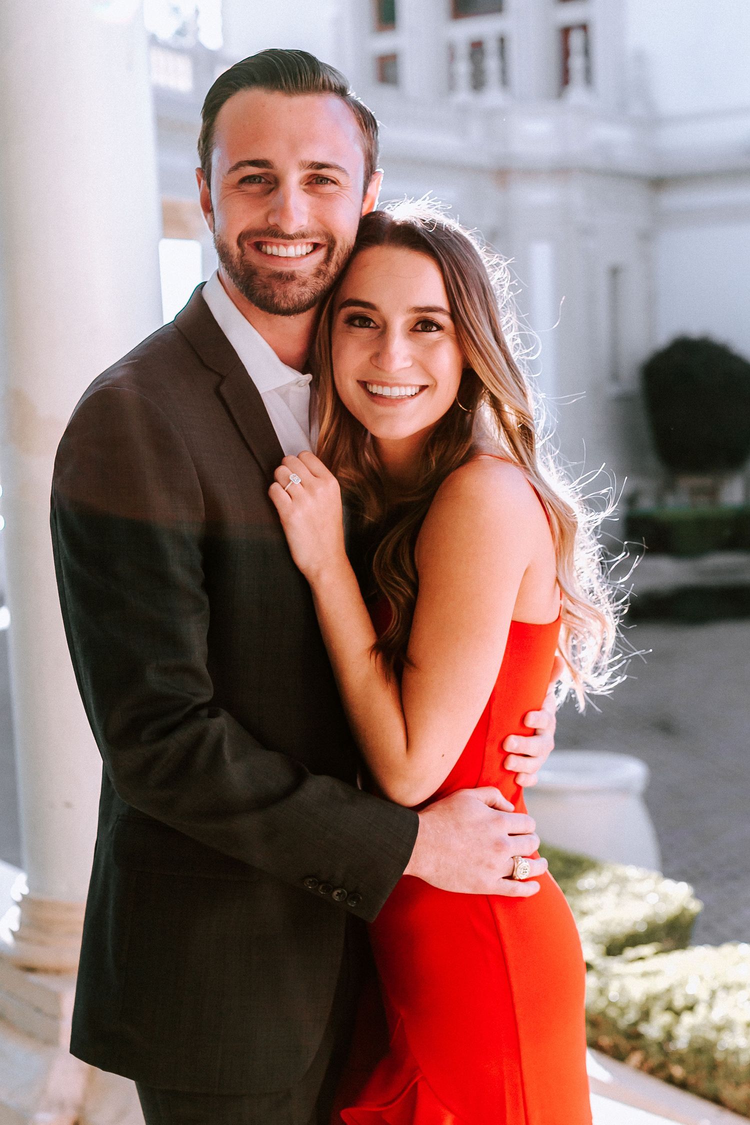 Happy couple hugging and smiling outdoors, woman in red dress and man in dark blazer.