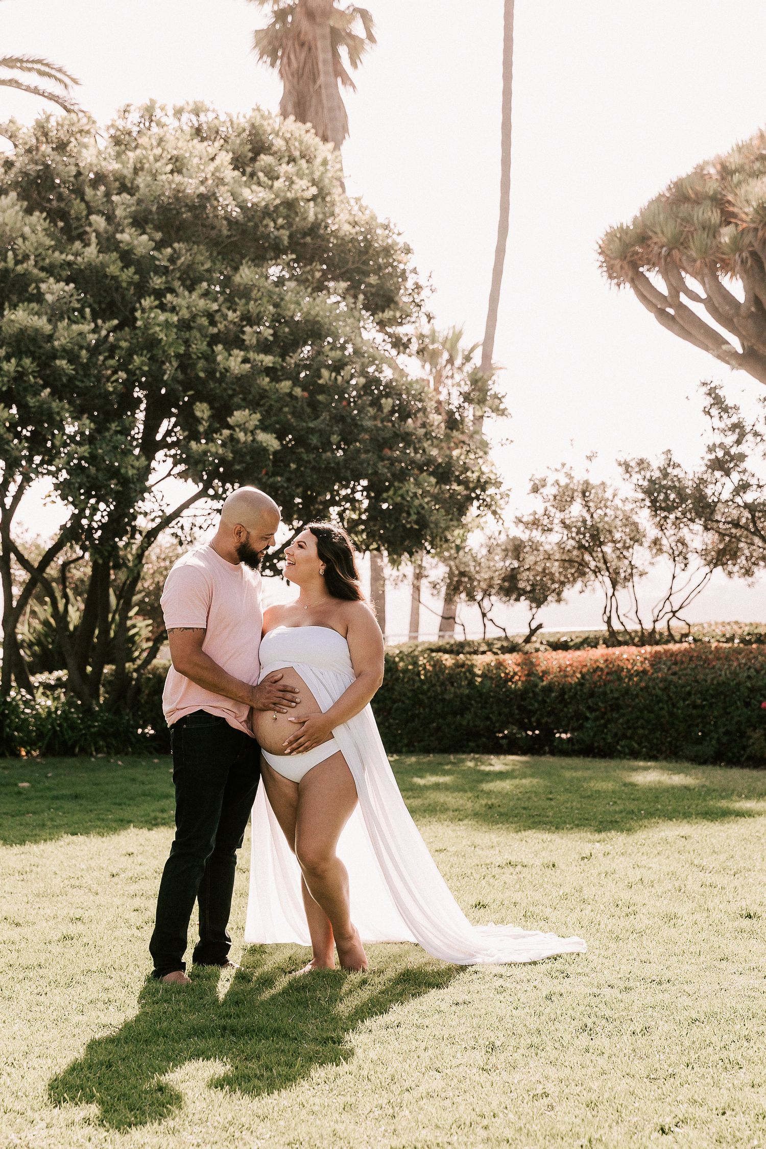 Expecting couple sharing a kiss in lush garden, woman in white maternity dress, palm trees behind.