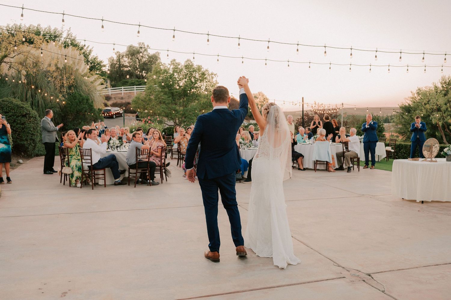 Bride and groom raise hands on outdoor wedding dance floor with string lights and seated guests at dusk.