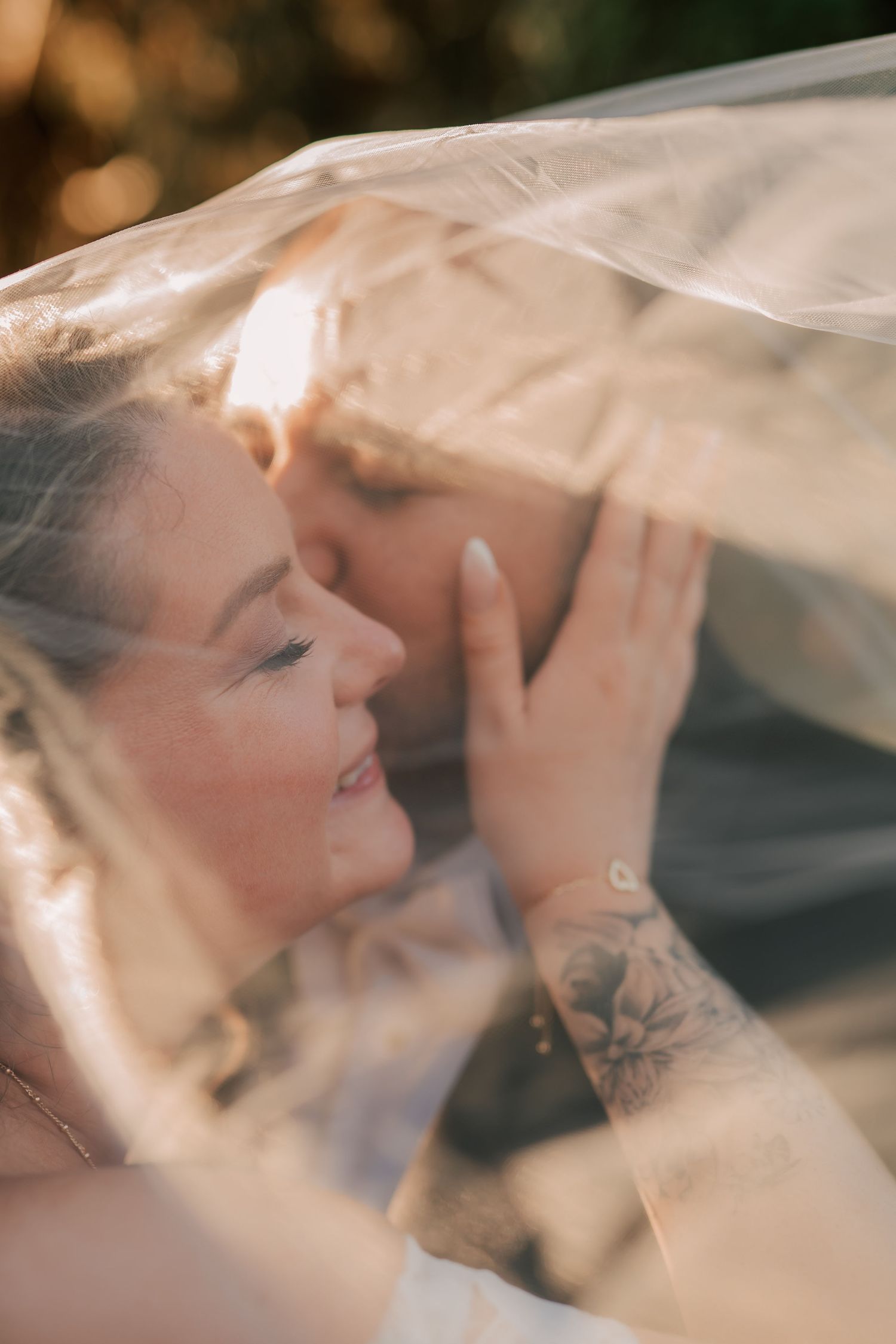 Bride and groom sharing a romantic moment under a wedding veil, with golden sunlight and a floral tattoo visible.