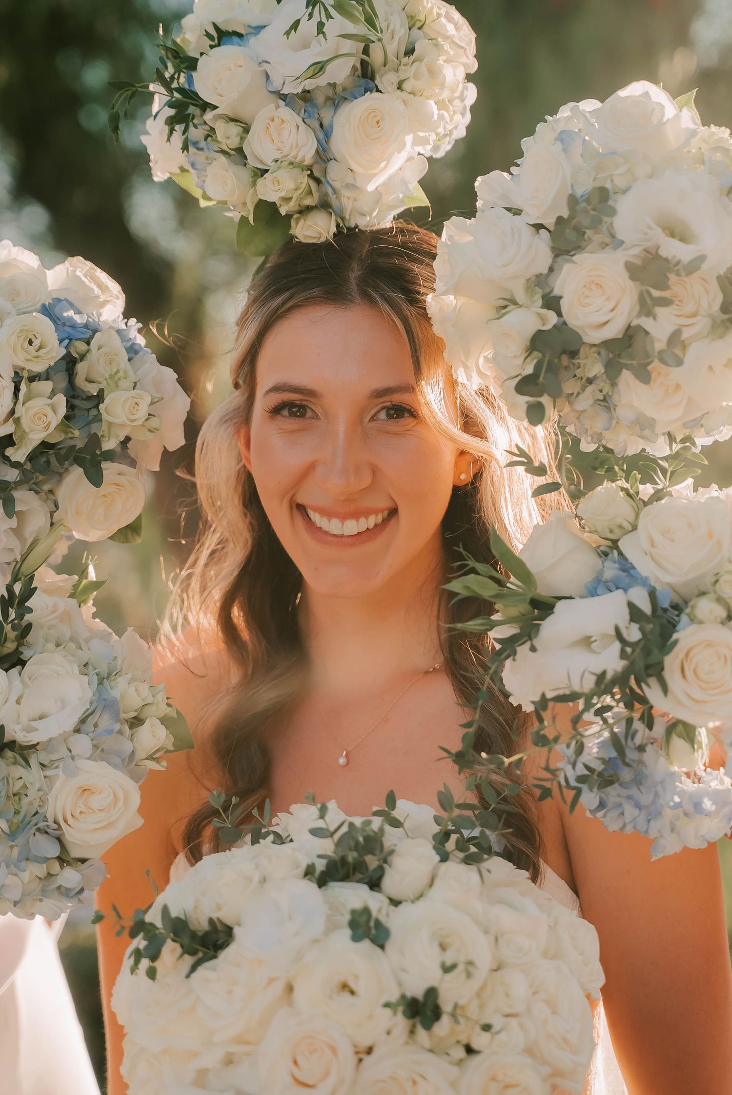 Smiling bride surrounded by lush white and blue floral arrangements, holding a bouquet of white roses outdoors.