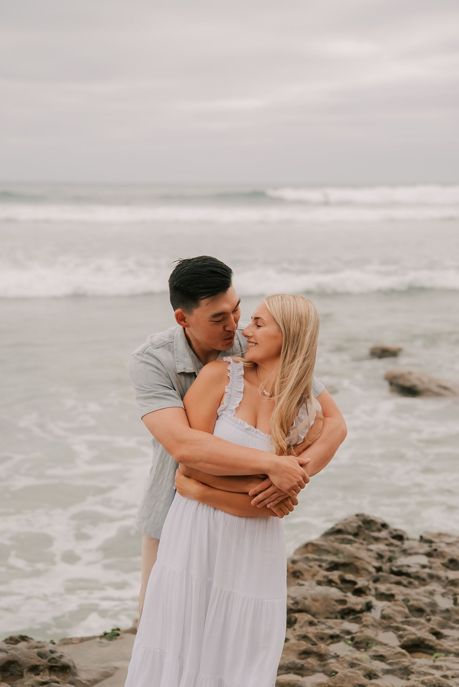 Couple embracing on a rocky beach at the shore, the man hugging a blonde woman in a white dress from behind under a soft overcast sky.