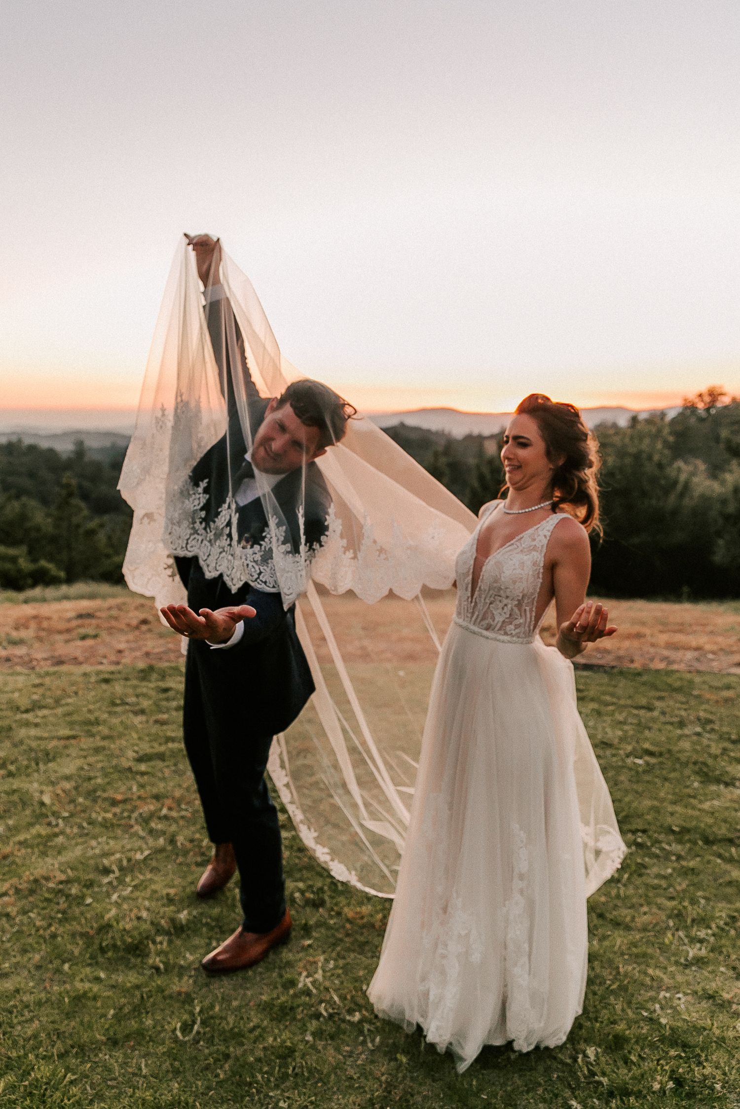 Bride and groom playfully lift lace wedding veil at sunset on a scenic hilltop during outdoor wedding photos.