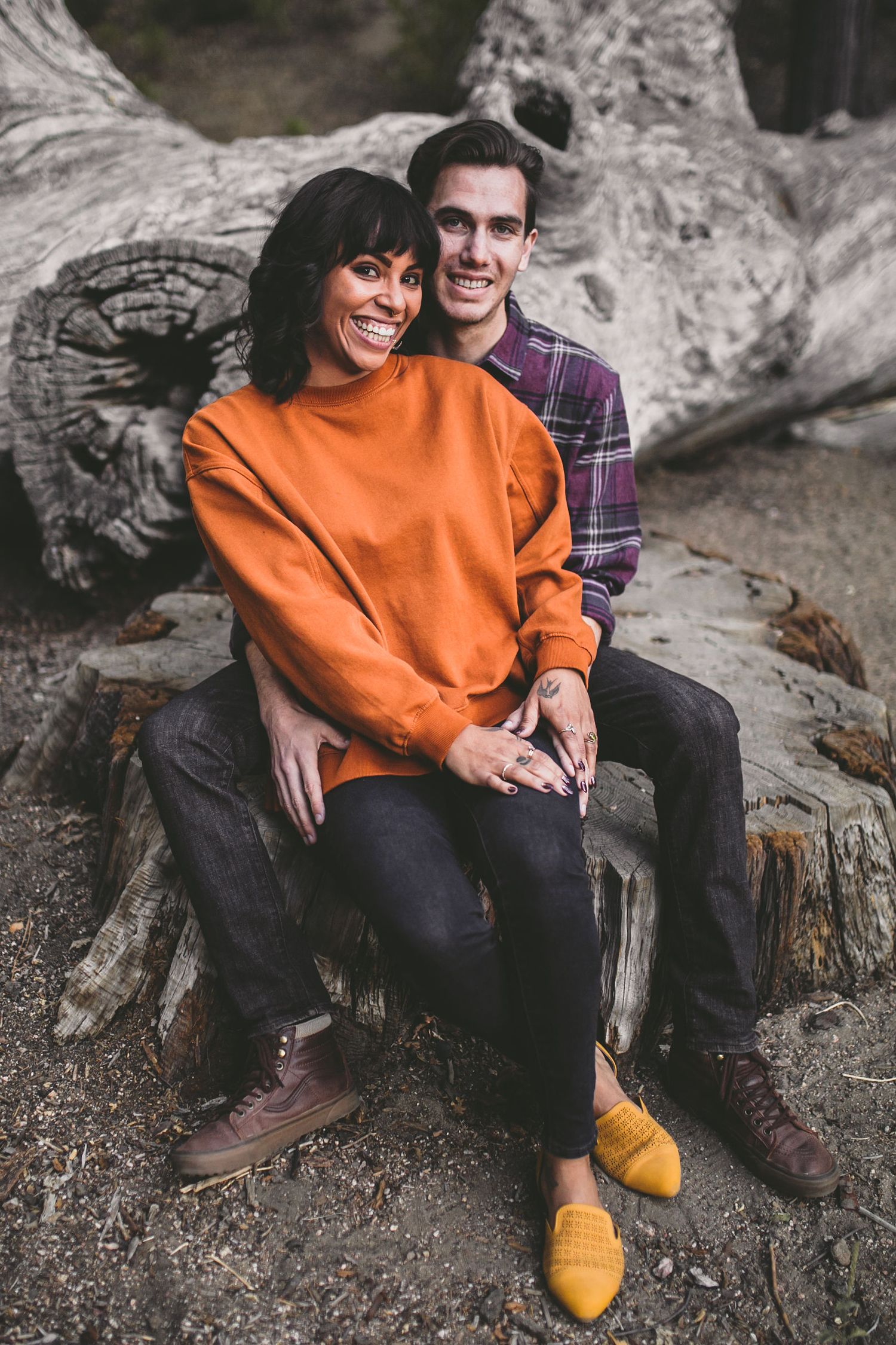 A smiling couple sits on a fallen log outdoors, woman in orange sweater and yellow shoes, man in plaid shirt.