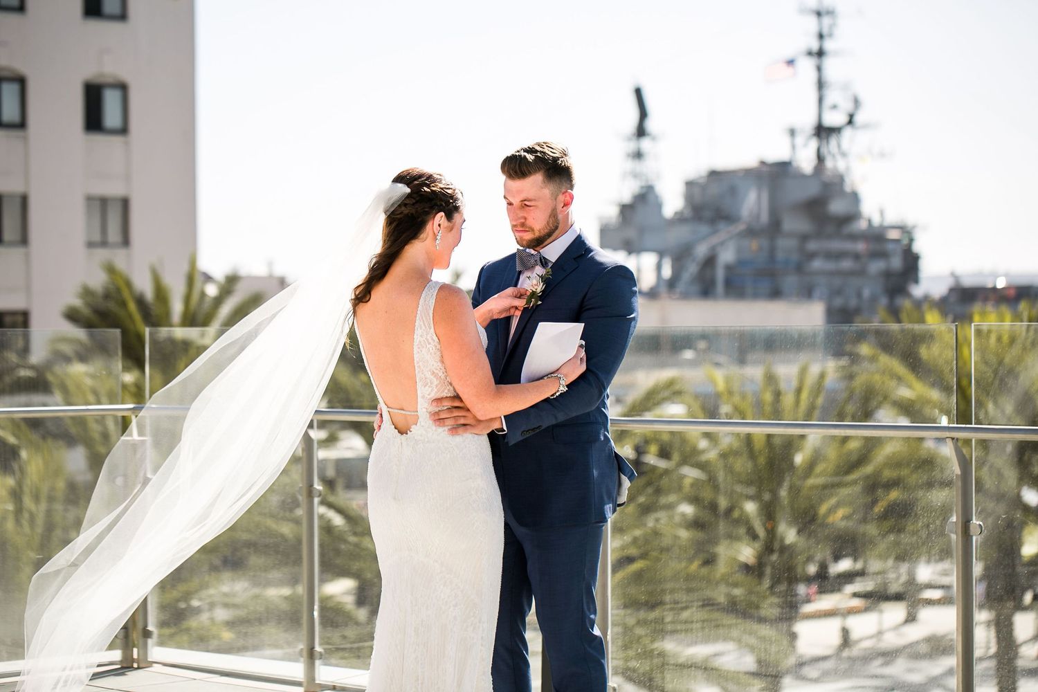 Groom reads vows to bride in white gown on rooftop terrace with navy ship and palm trees in background.