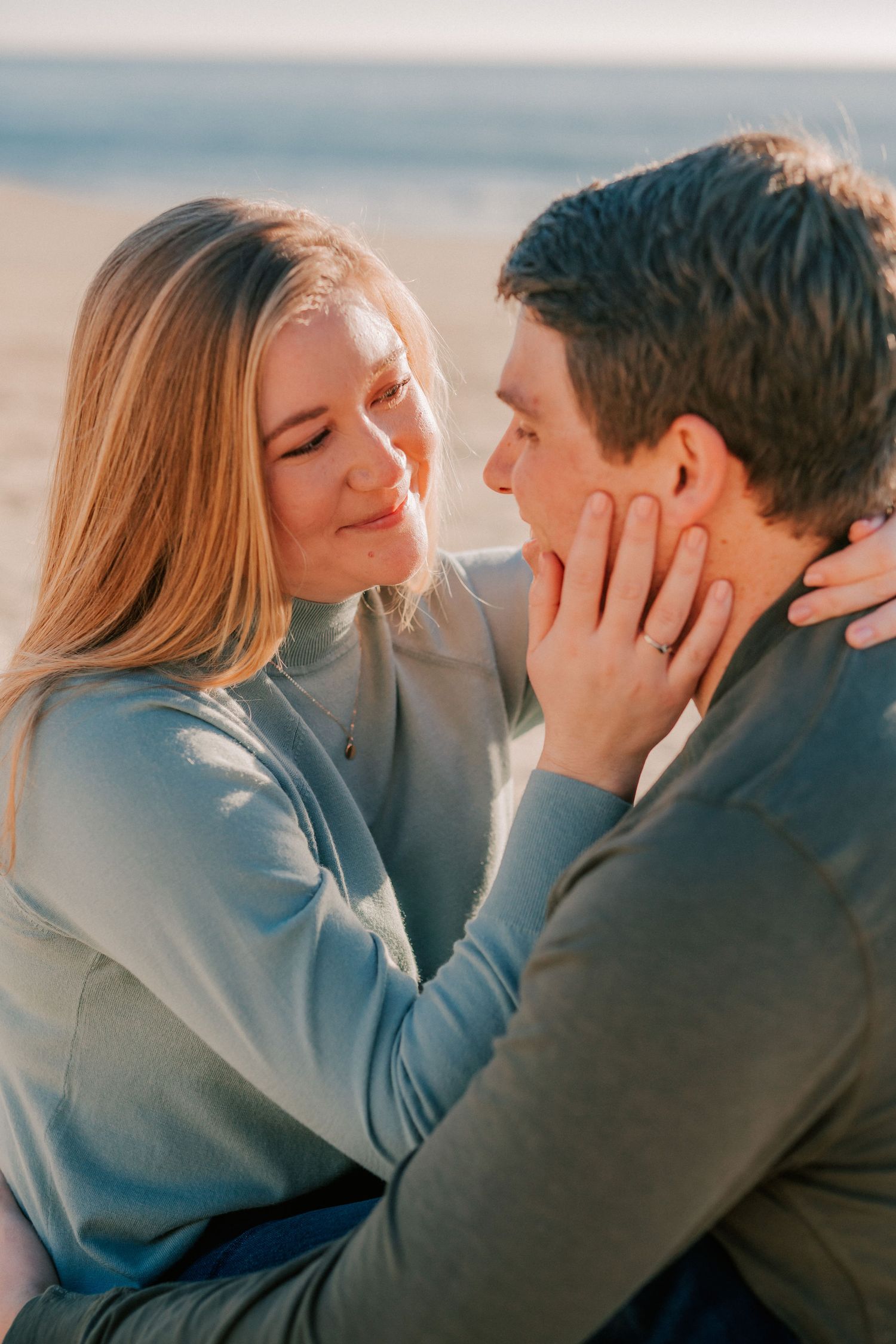 A couple shares a tender moment on the beach, with the woman gently cupping the man's face while smiling warmly.