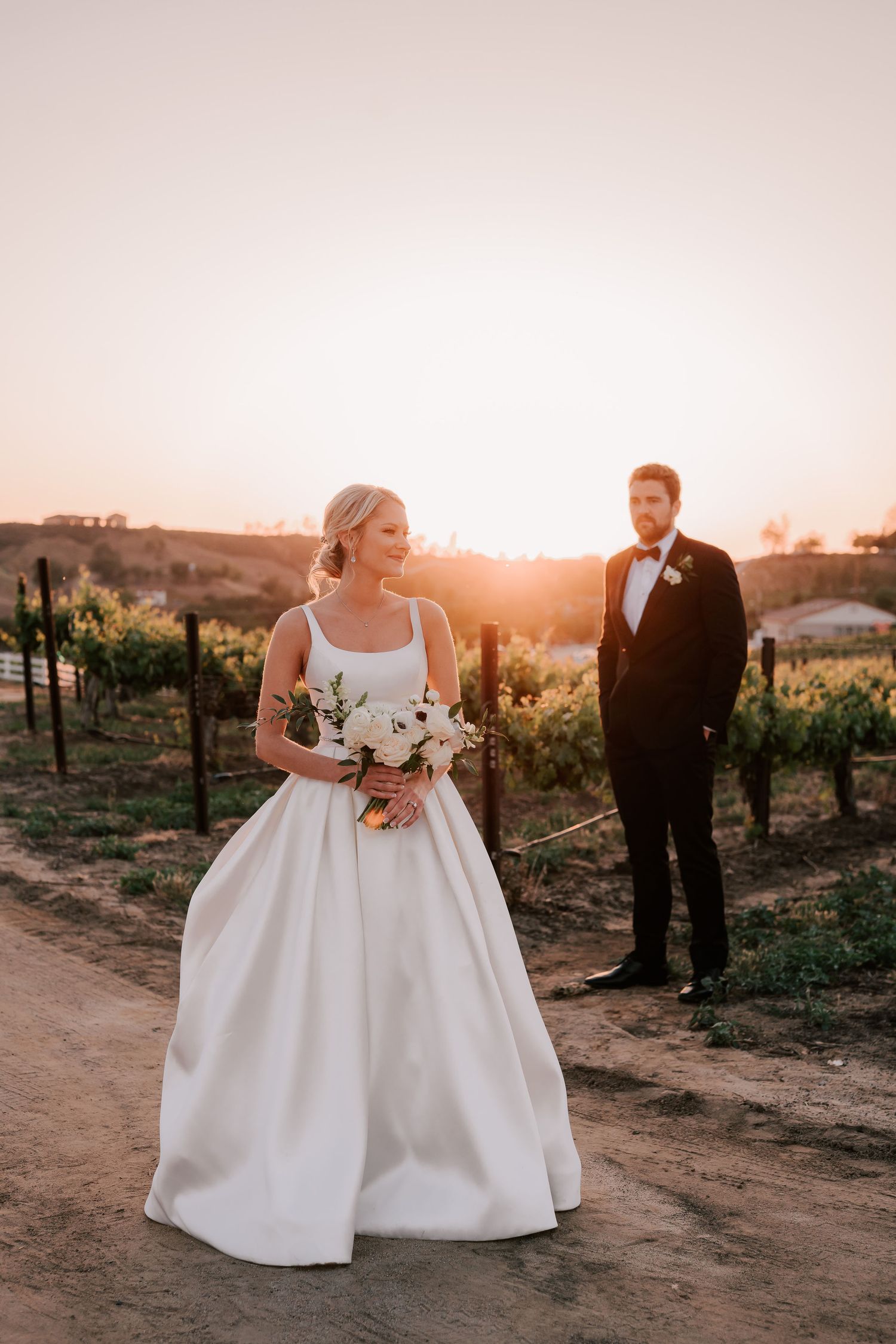 Bride in white gown holds bouquet at sunset vineyard wedding, groom in tuxedo stands behind her.