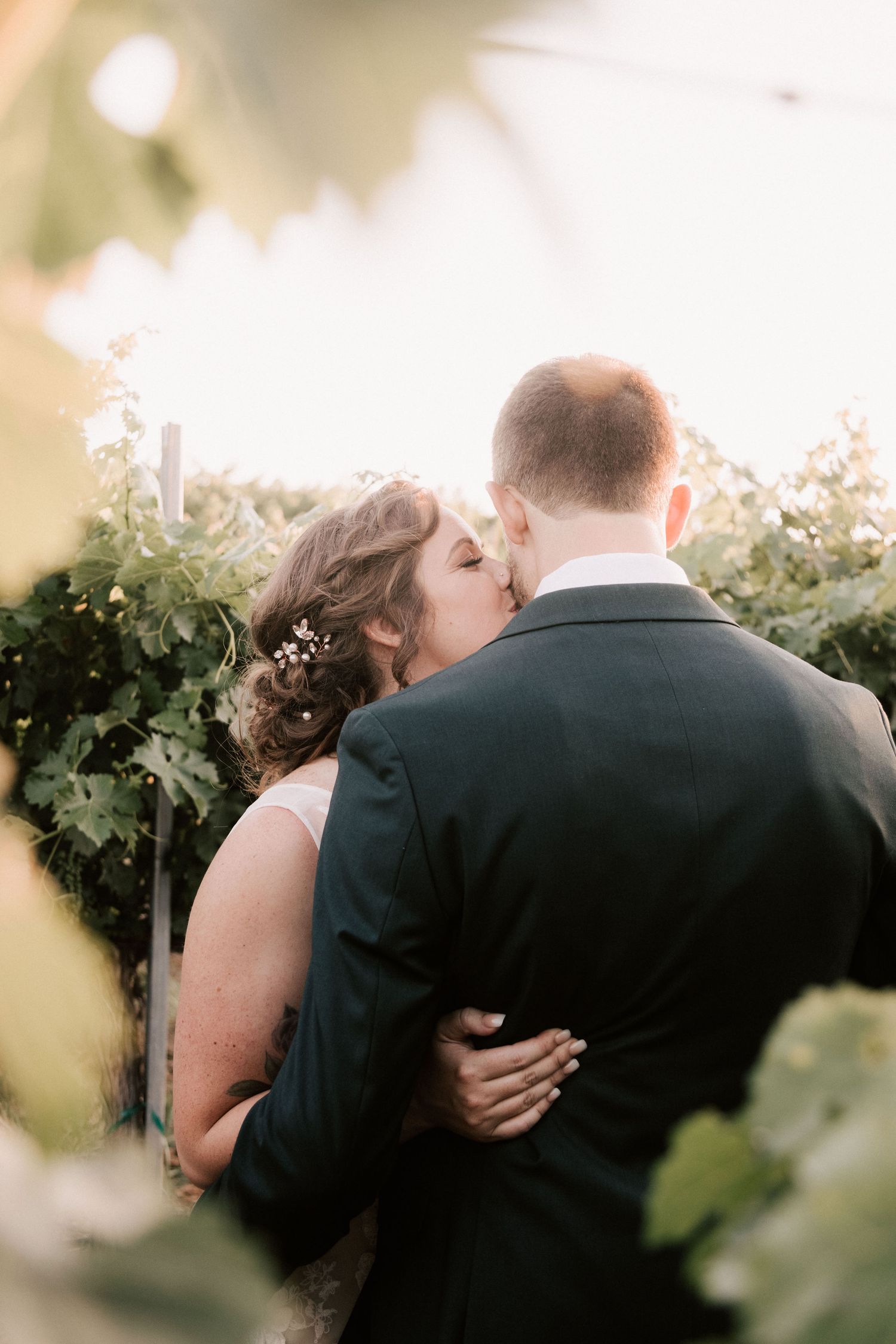 A bride and groom share a romantic kiss surrounded by lush green vineyard foliage during their wedding.