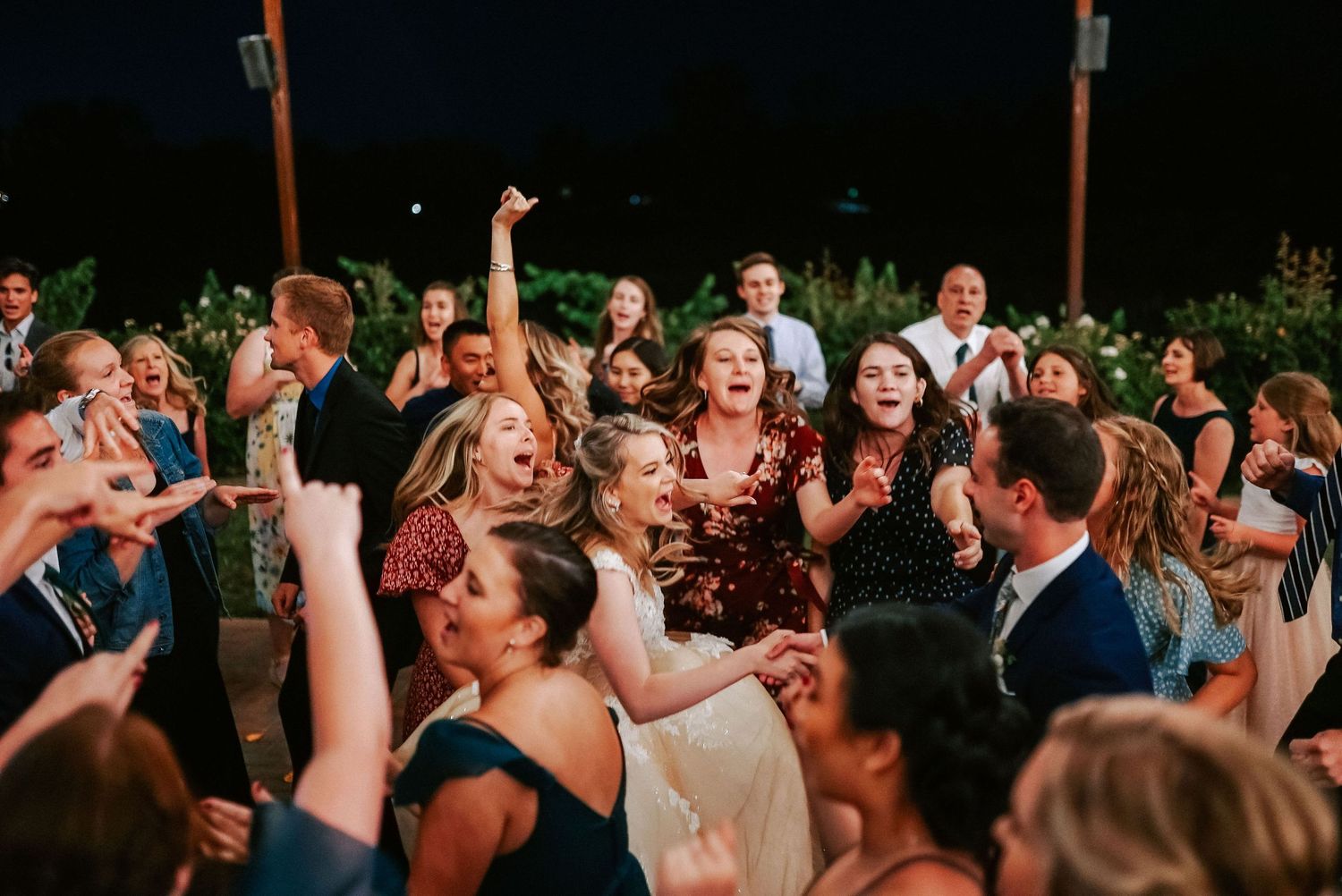 Joyful wedding reception guests dancing and celebrating outdoors at night, with bride in white dress surrounded by friends.