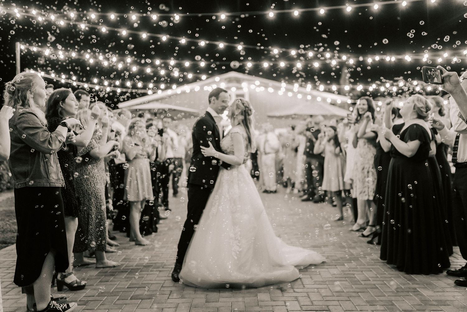 Black and white wedding photo of couple kissing under string lights with guests blowing bubbles outdoors.