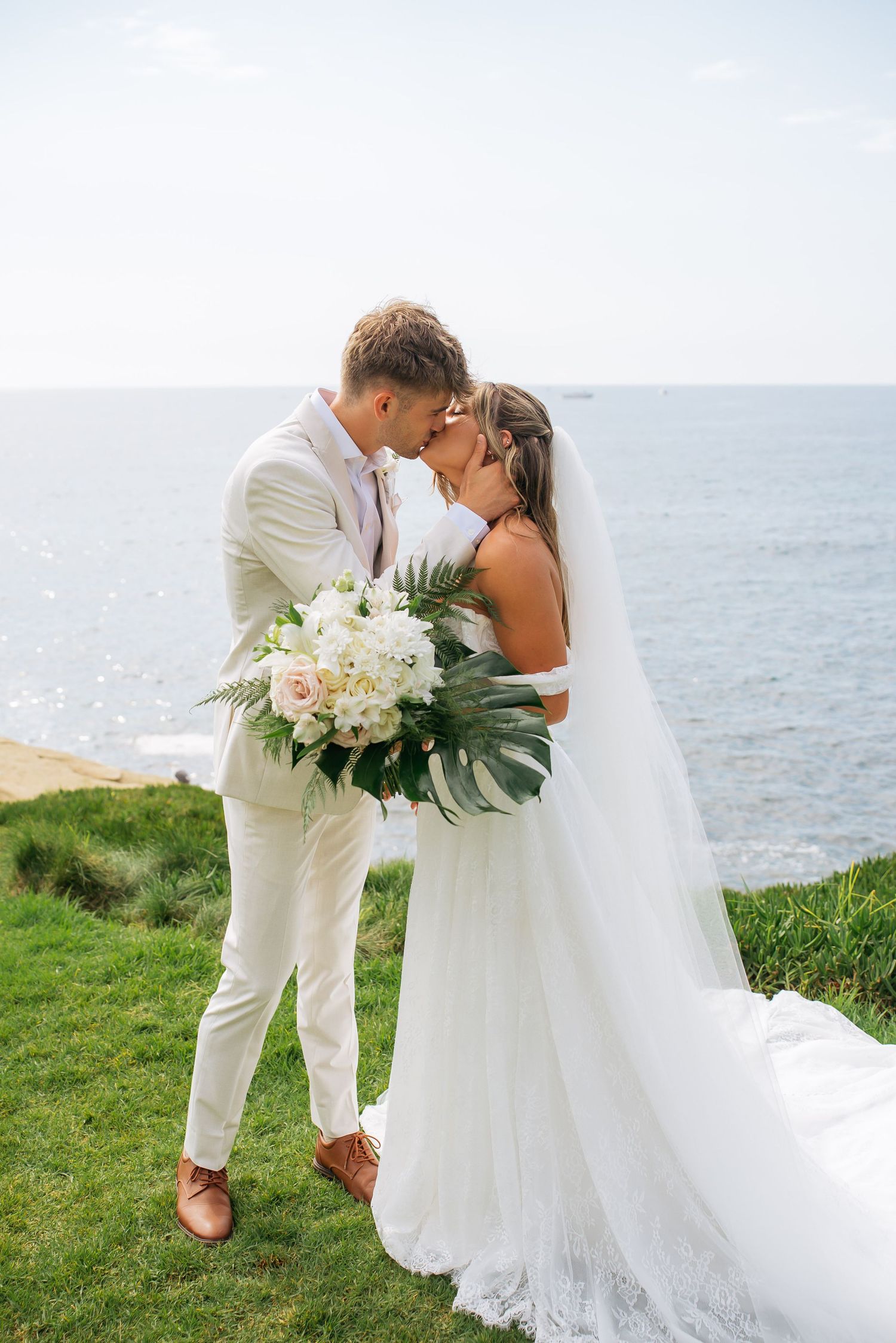 Bride and groom share a romantic kiss overlooking the ocean, bride holding a white floral bouquet with tropical leaves.
