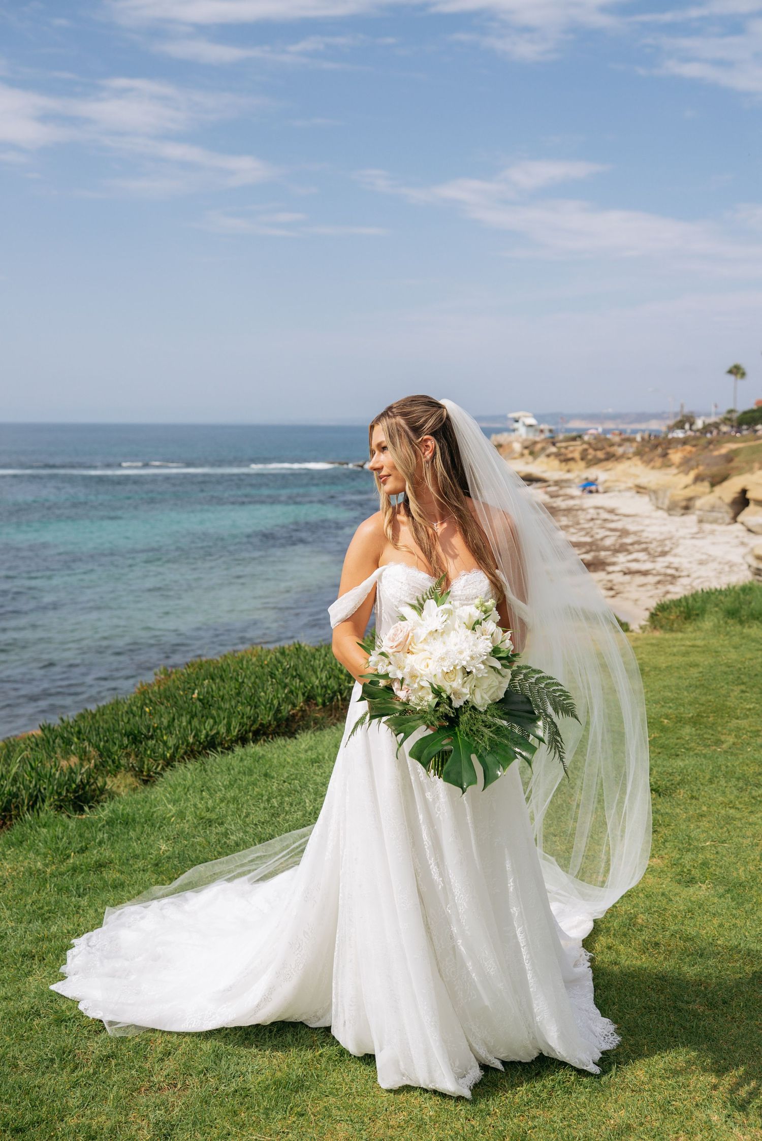 Bride in off-shoulder gown with veil holds white bouquet by ocean cliffside on sunny wedding day.