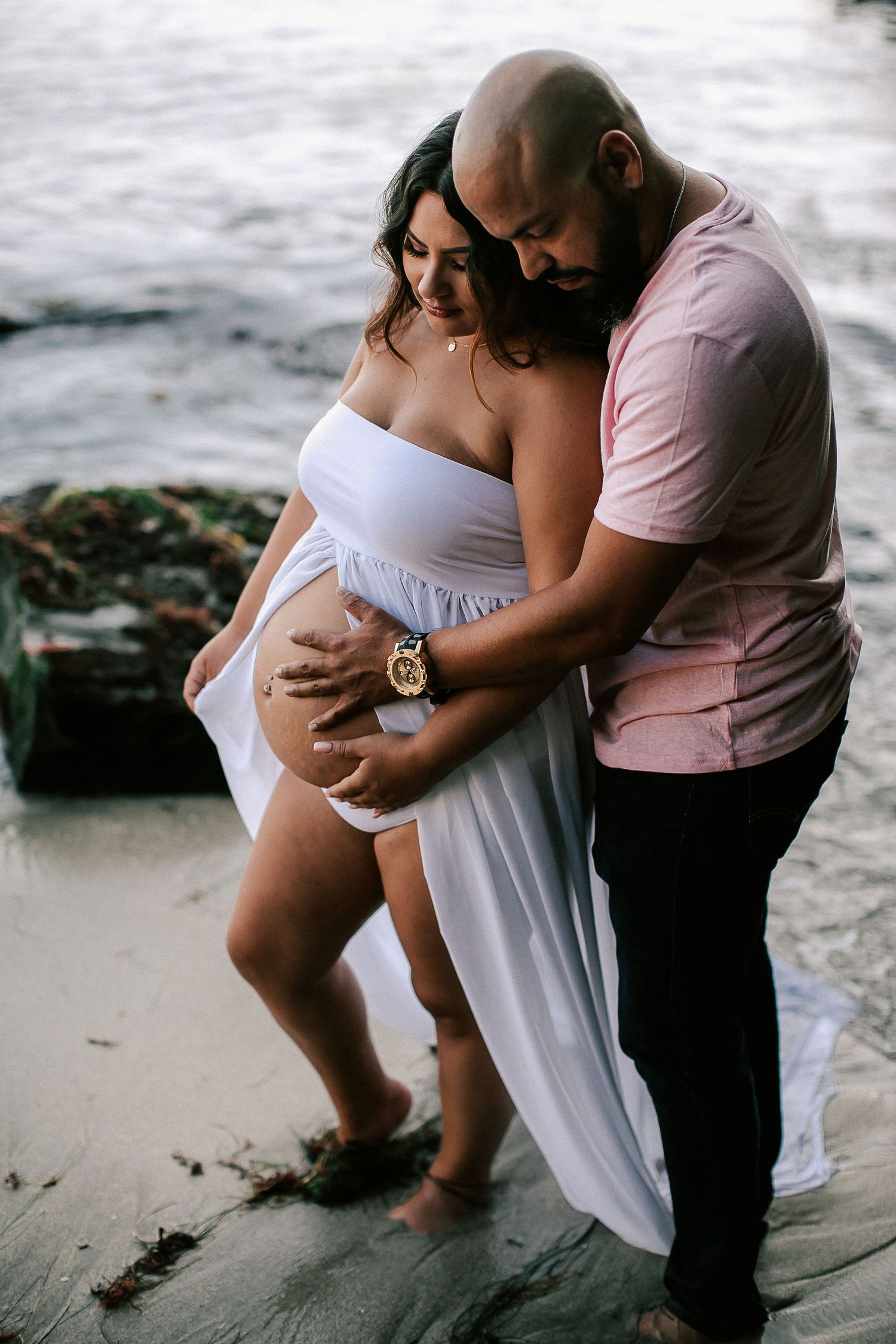 Man embracing pregnant woman in white dress on beach, both looking down at her baby bump.