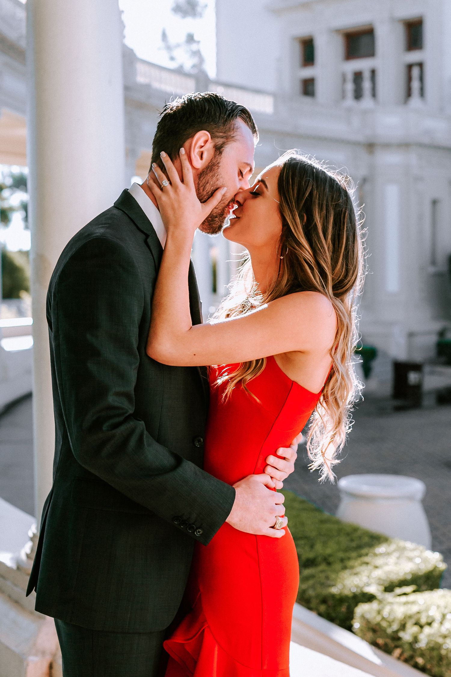 Couple in formal attire sharing a kiss outdoors, woman in red dress embracing man in dark suit.