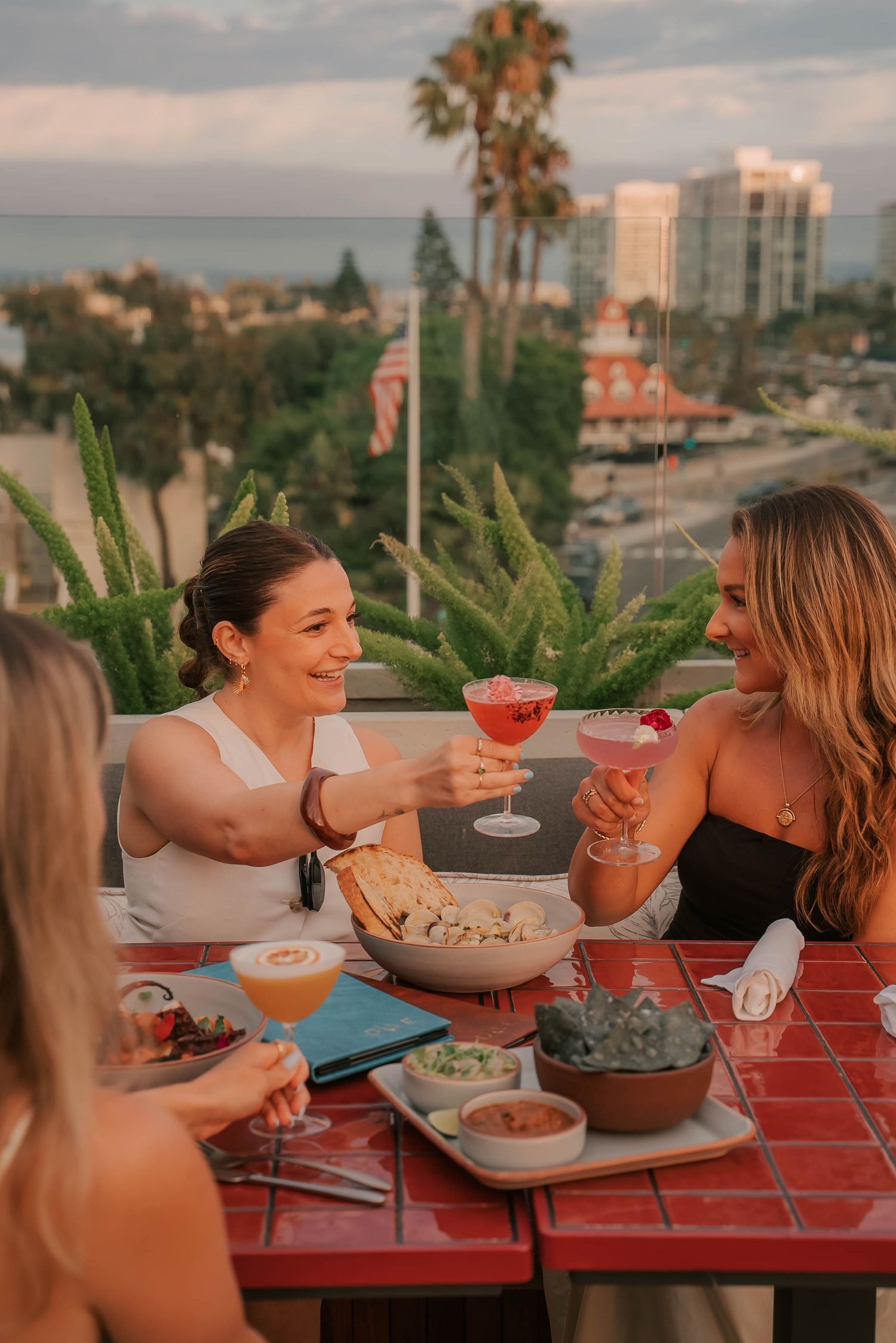 Three women toasting with colorful cocktails on a rooftop patio with palm trees and ocean views in the background.