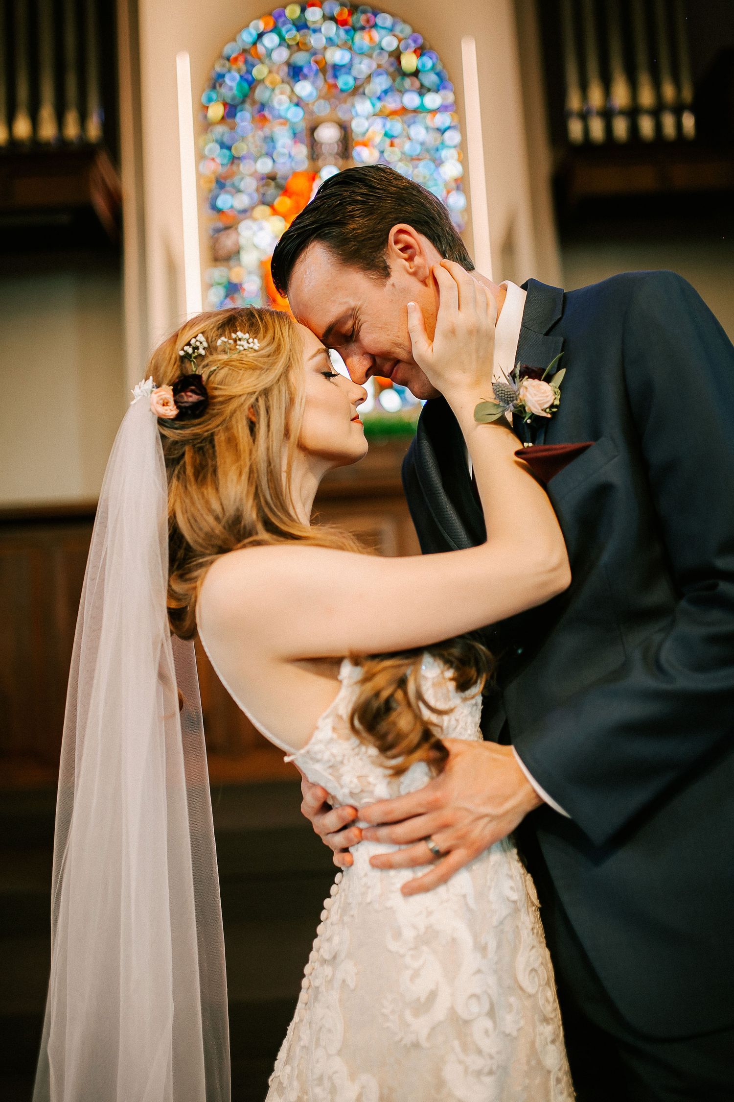 Bride and groom share a romantic kiss at church altar with colorful stained glass window backdrop.