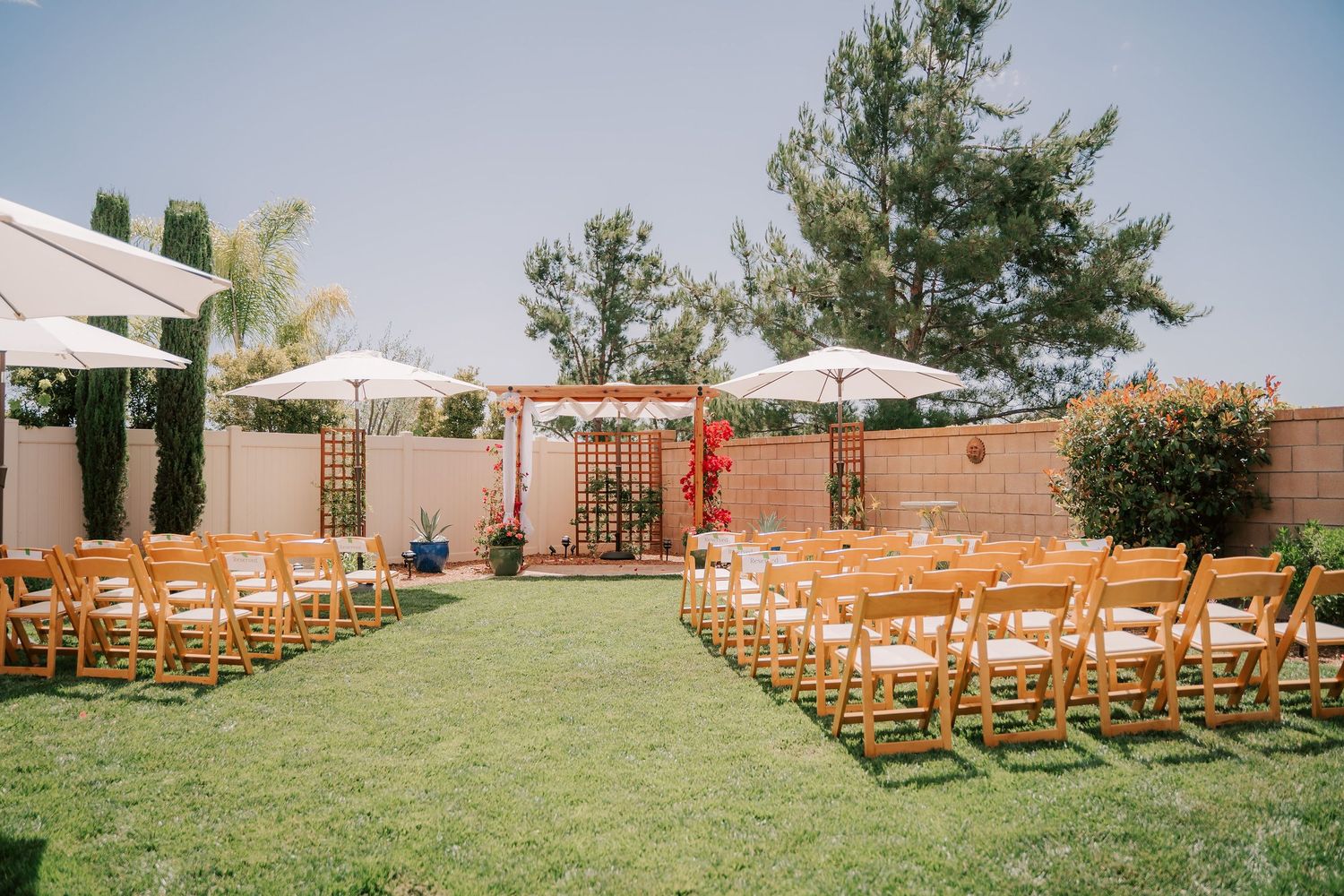 Outdoor wedding ceremony setup with wooden chairs on green lawn, white umbrellas, and floral arch in sunny backyard venue.