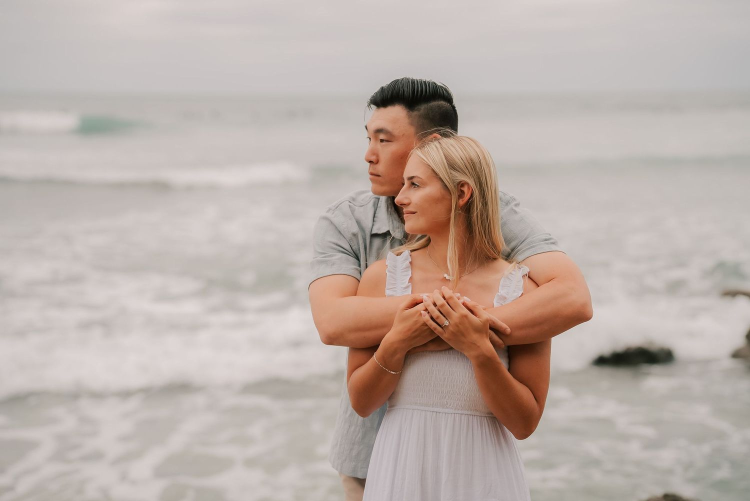 Couple embracing on a beach, man hugging woman in white dress with ocean waves in background.