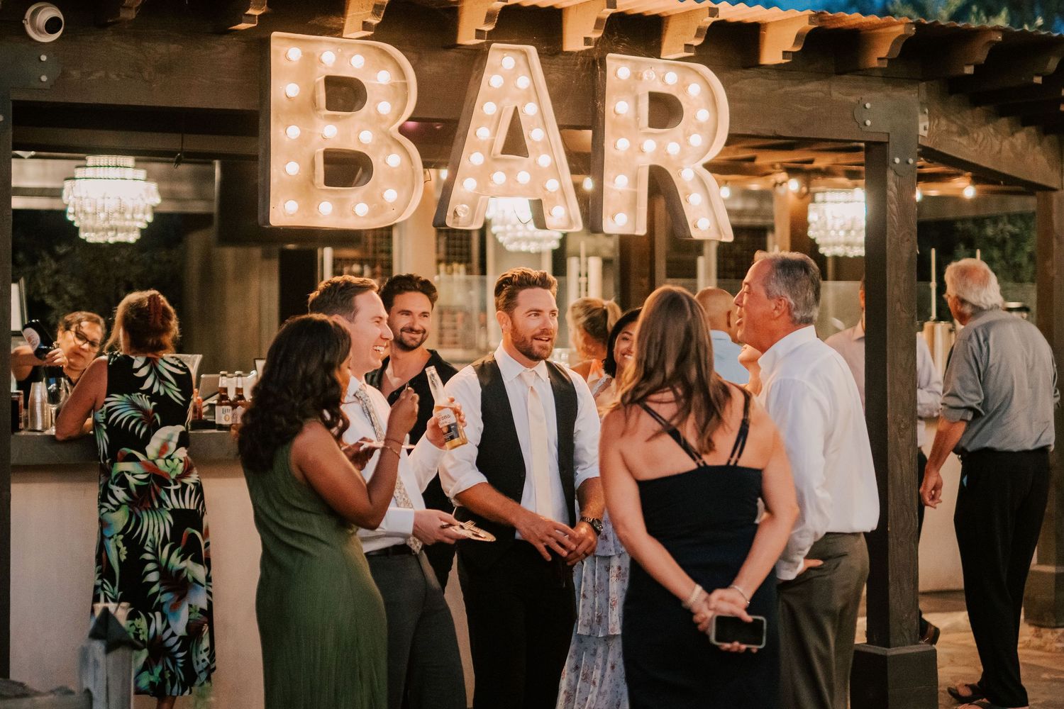 Wedding guests socializing near a lit BAR sign at an outdoor evening reception venue.