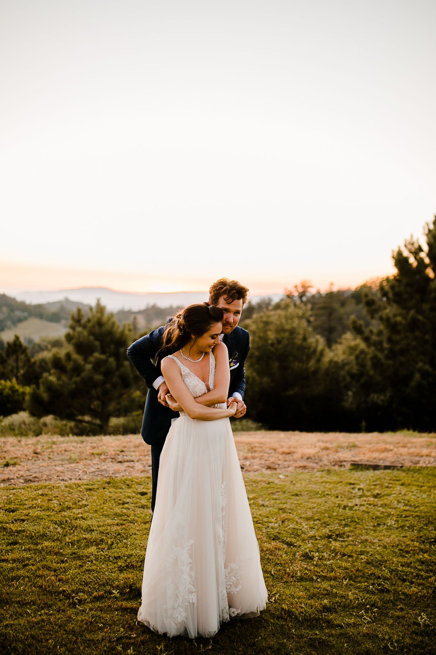 A couple shares a romantic embrace at golden hour, with the groom hugging the bride in her lace wedding gown outdoors.