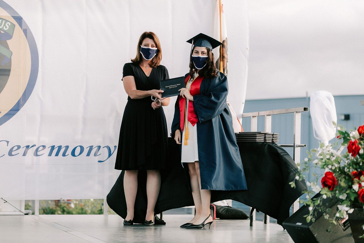 Two women in face masks at a graduation ceremony, one in cap and gown receiving a handshake on stage.