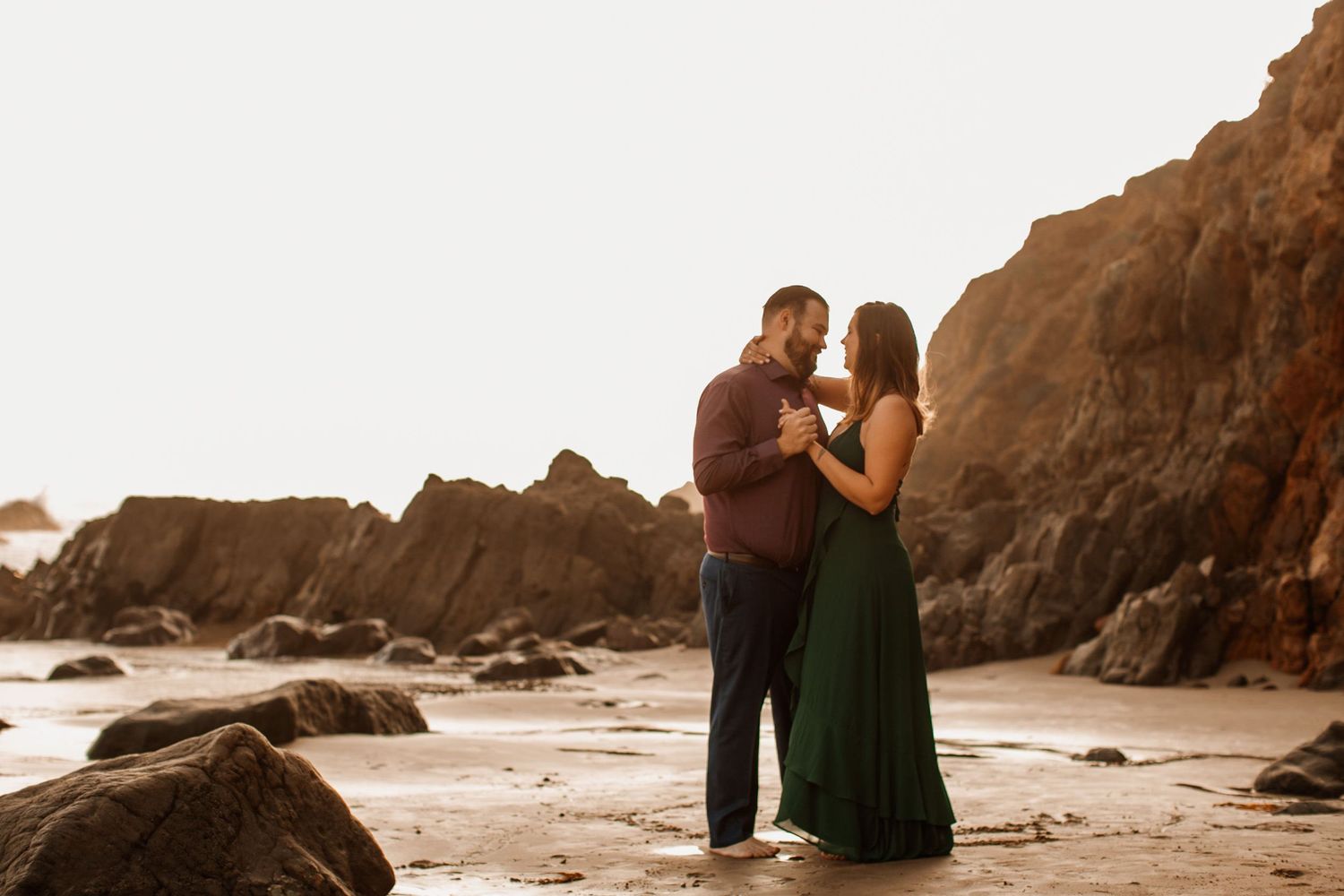 Couple embracing romantically on a rocky beach at golden hour, woman in green dress, man in burgundy shirt.