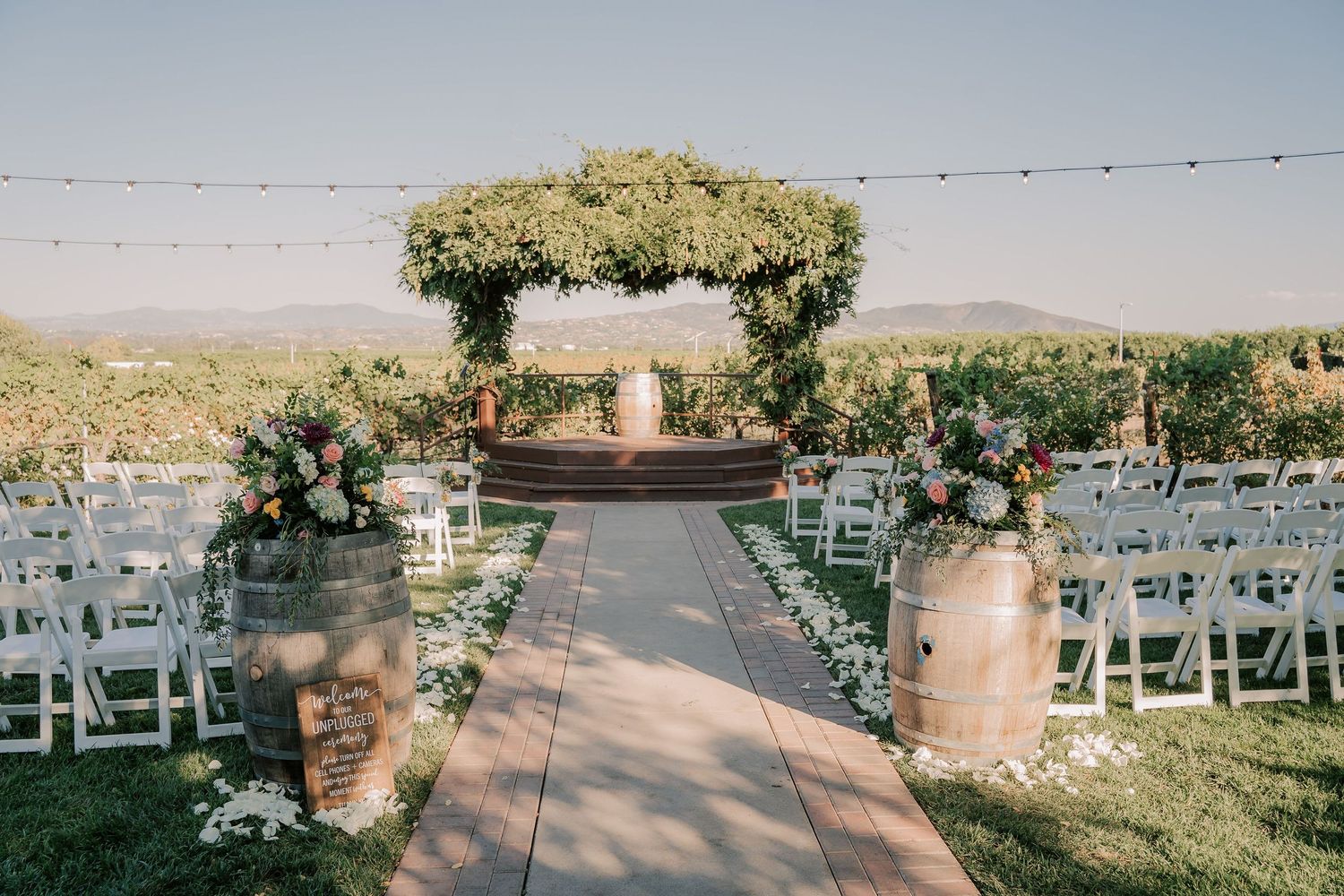Elegant outdoor wedding ceremony aisle with floral arrangements, wine barrels, white chairs, and a vine-covered arbor at a vineyard.
