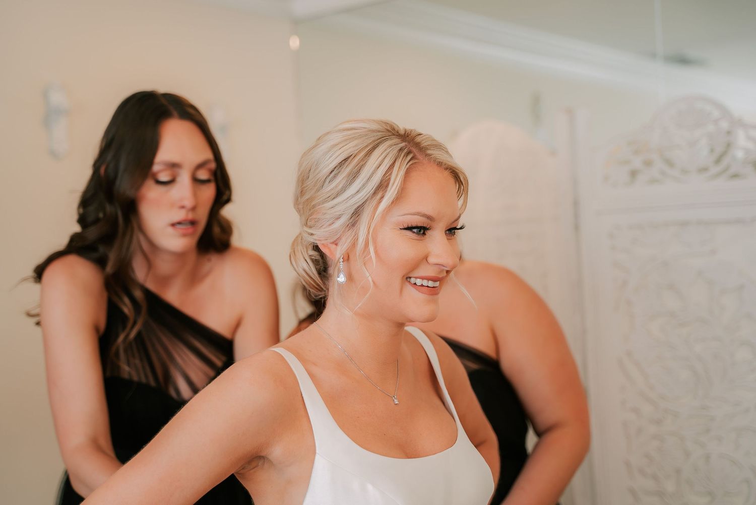 Bridesmaid in black dress helping bride in white gown get ready, smiling warmly in bright dressing room.