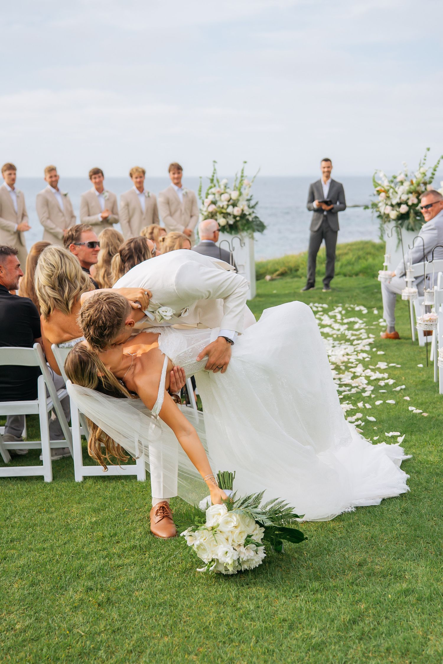 Groom dips bride in dramatic kiss at outdoor oceanfront wedding ceremony on green lawn.