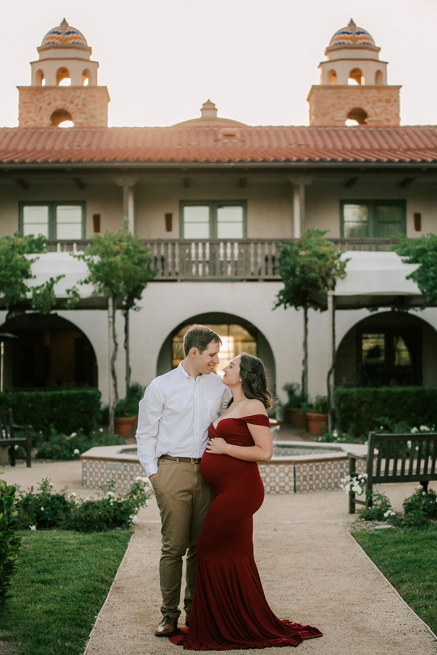 Couple poses for maternity photo in red gown before Spanish-style building with courtyard fountain.