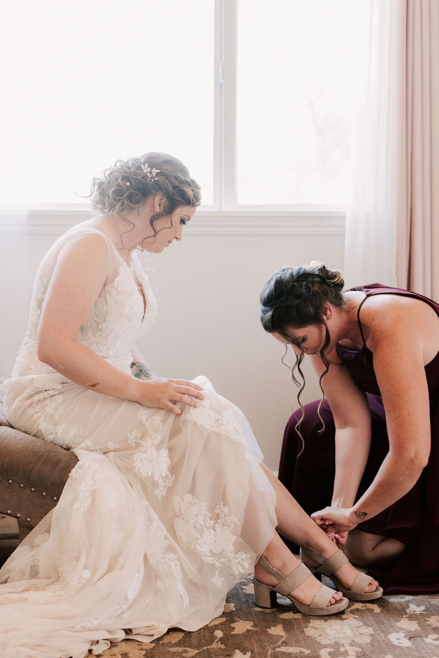 Bridesmaid in purple dress helps bride in lace wedding gown put on strappy heels while seated near bright window.