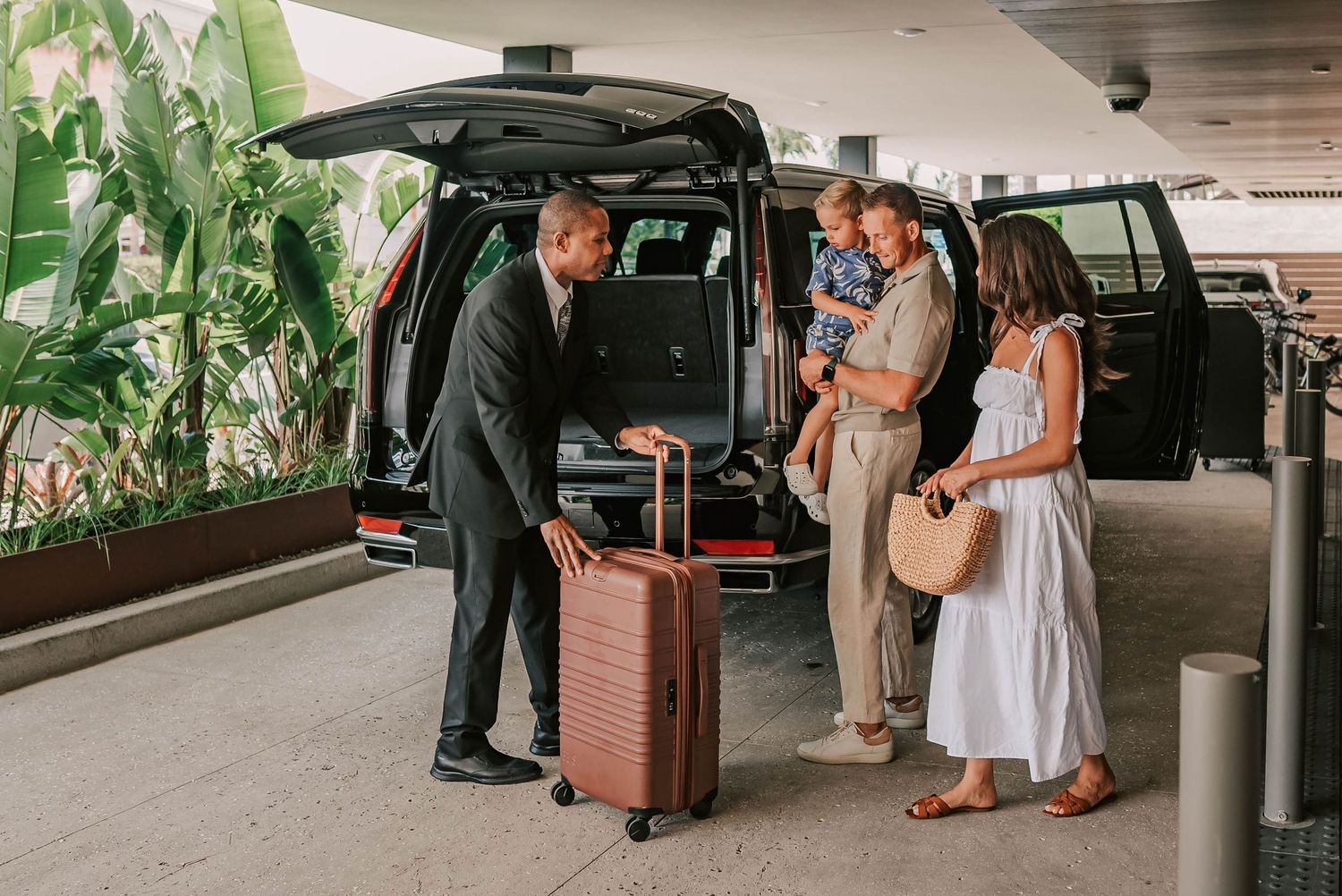 A chauffeur in a suit loads luggage into a black van for a family at a hotel or airport drop-off area.