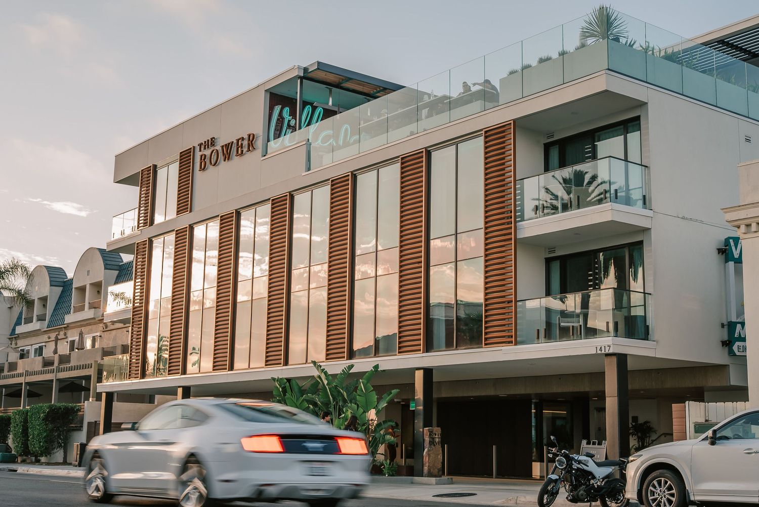 Modern multi-story hotel building with wood-paneled facade, rooftop terrace, and passing car on a sunny street.