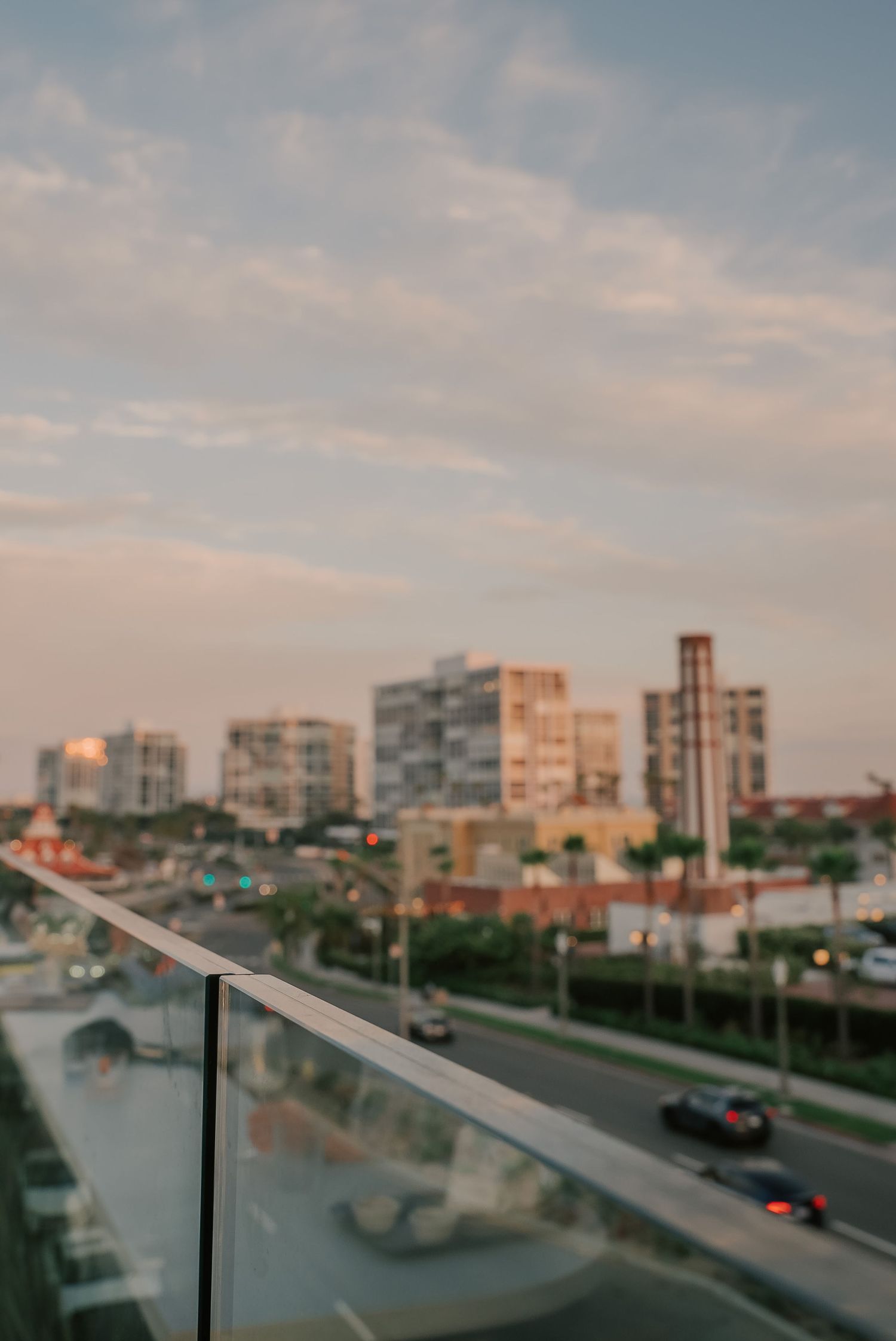 Blurry cityscape view from a glass balcony railing at sunset with buildings and traffic below.