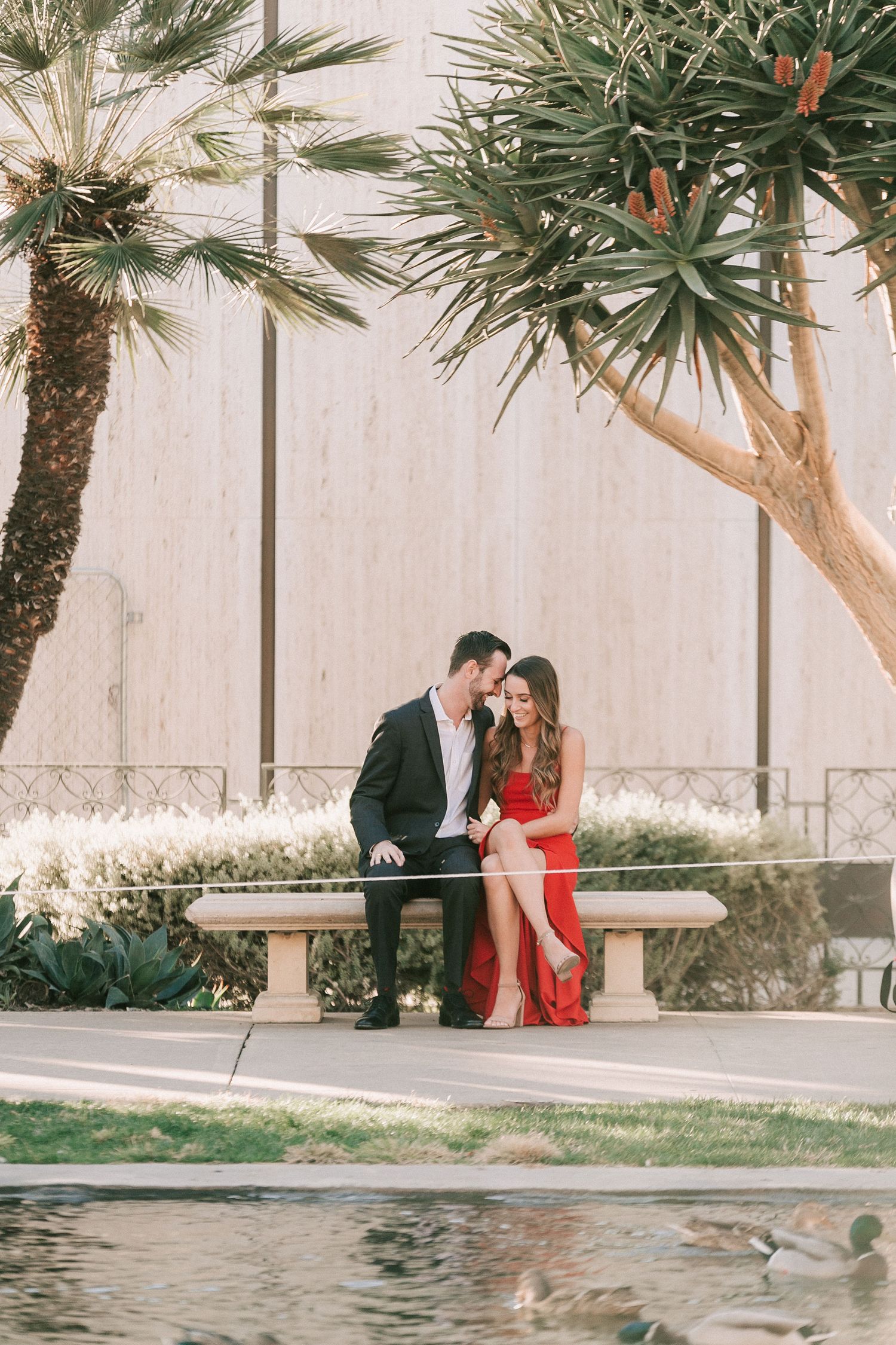 Couple sitting closely on a stone bench surrounded by palm trees and a reflecting pool outdoors.