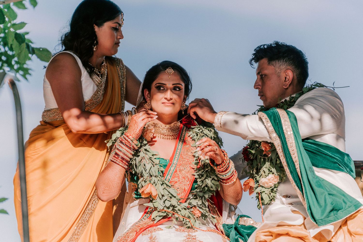 Bride being adorned with floral garland by attendants during traditional Indian wedding ceremony outdoors.