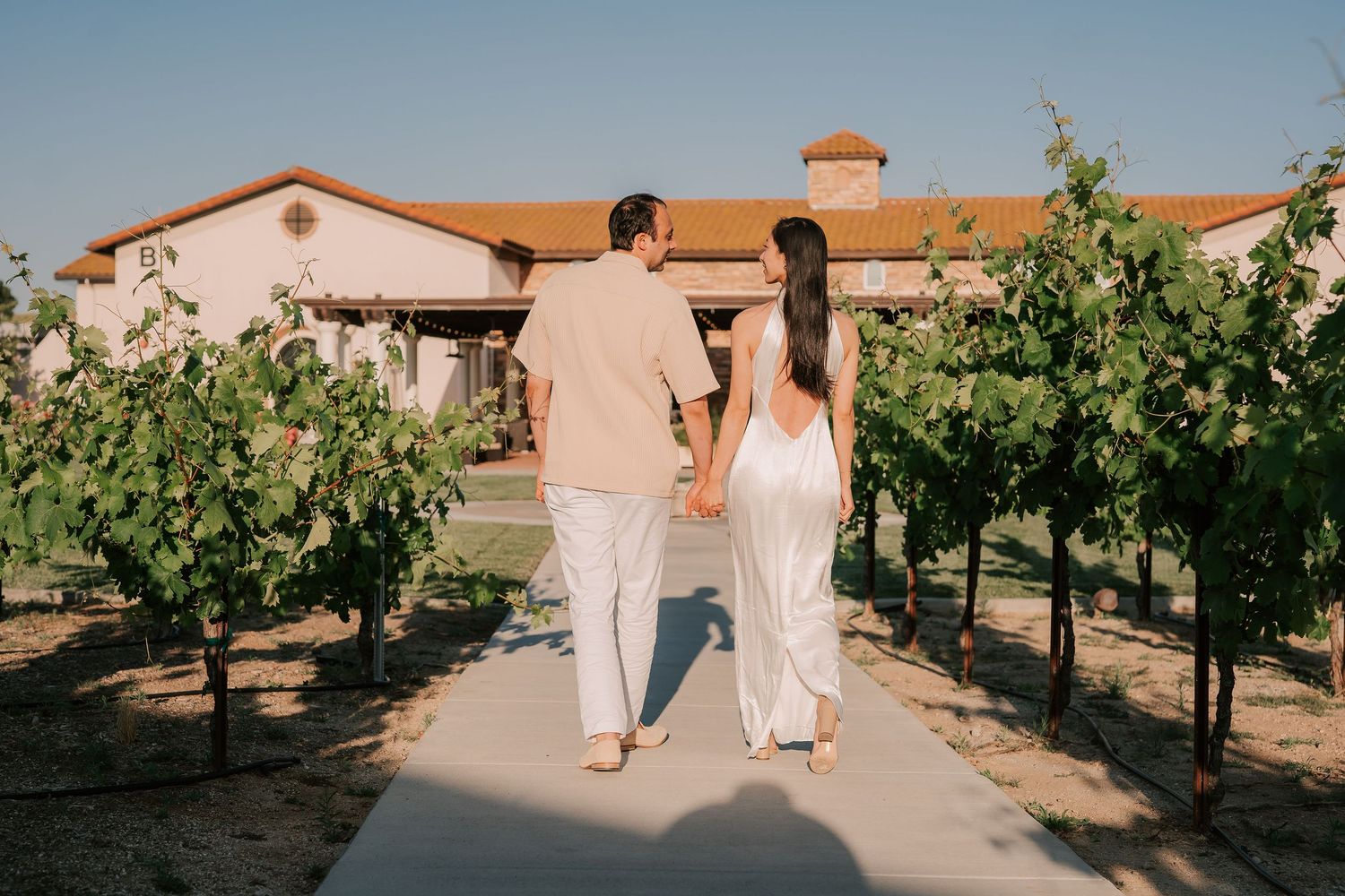 Couple walking hand in hand through a vineyard pathway toward a white winery building at golden hour.