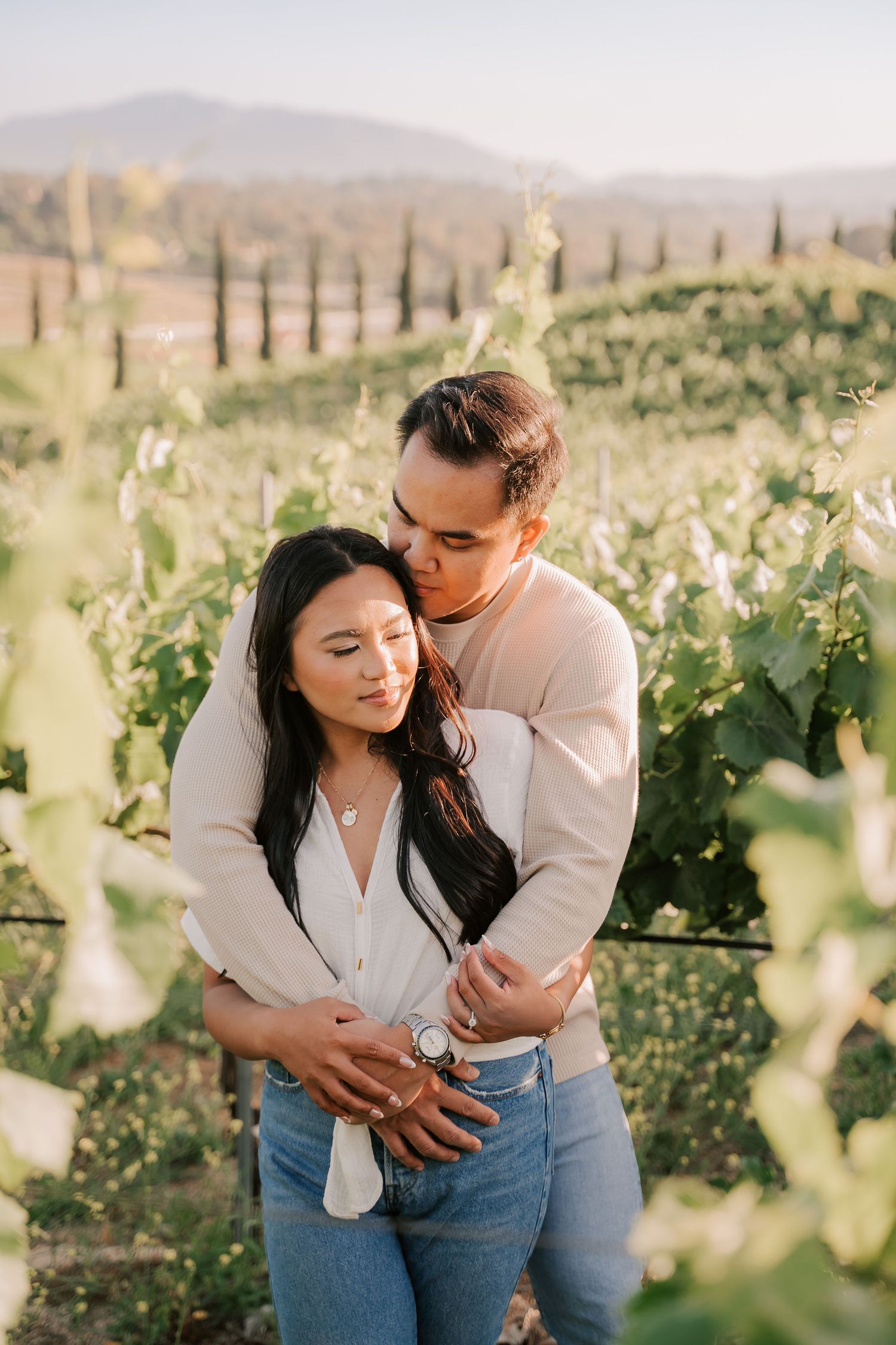 Couple embracing in a vineyard, man kissing woman's head, both wearing neutral tones and jeans, with rolling hills behind.