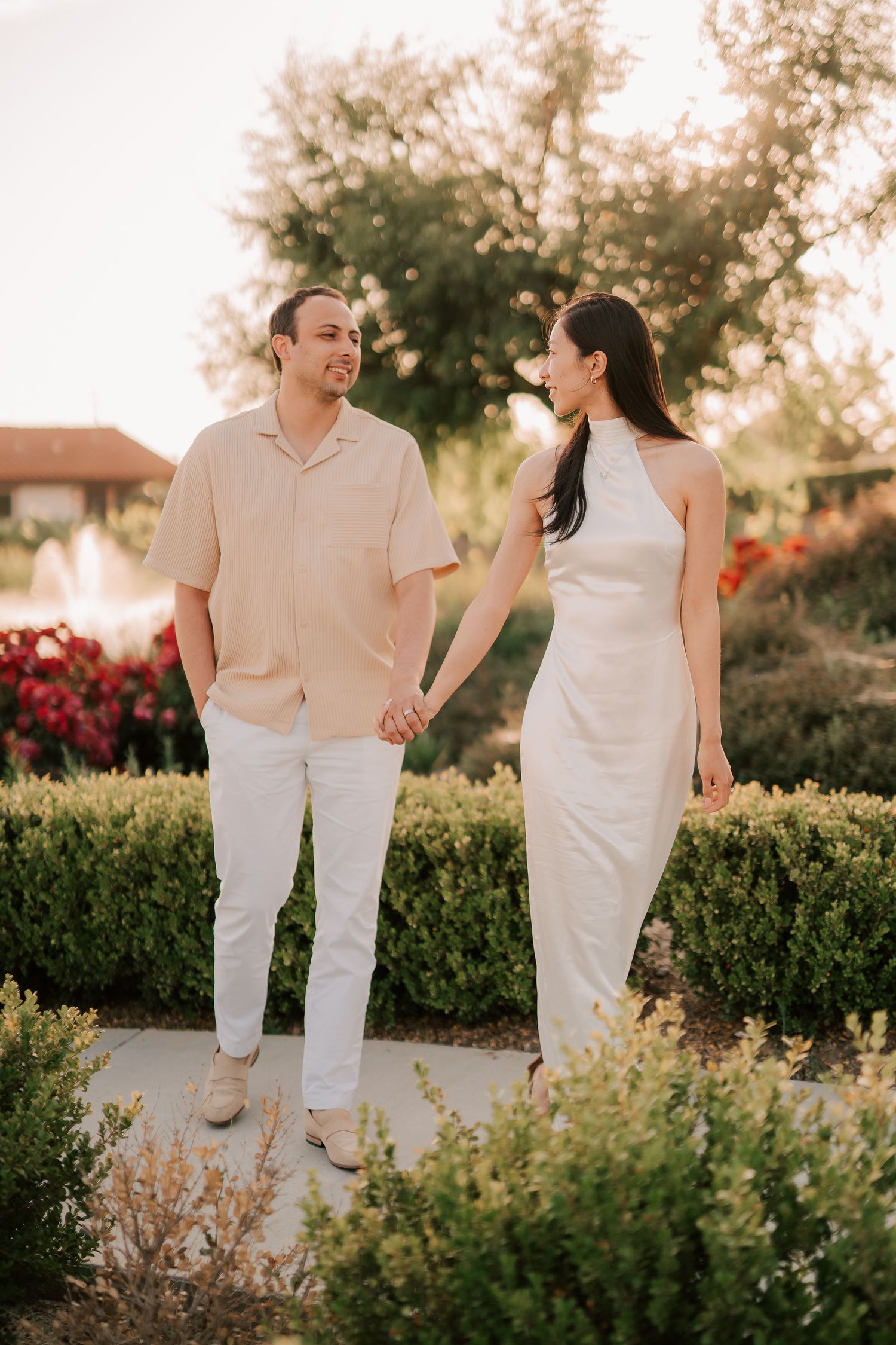 Couple holding hands and walking through a lush garden path at golden hour, man in beige shirt and woman in white dress.