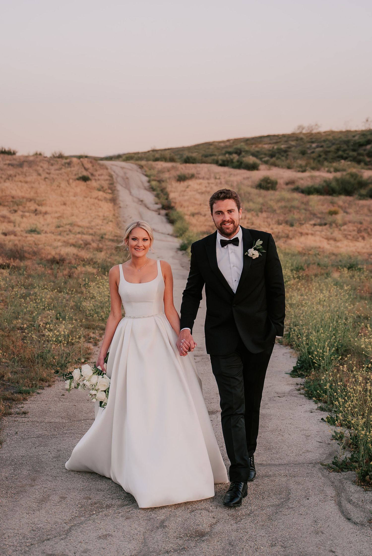 Bride in white gown and groom in black tuxedo hold hands walking down a dirt path at sunset.