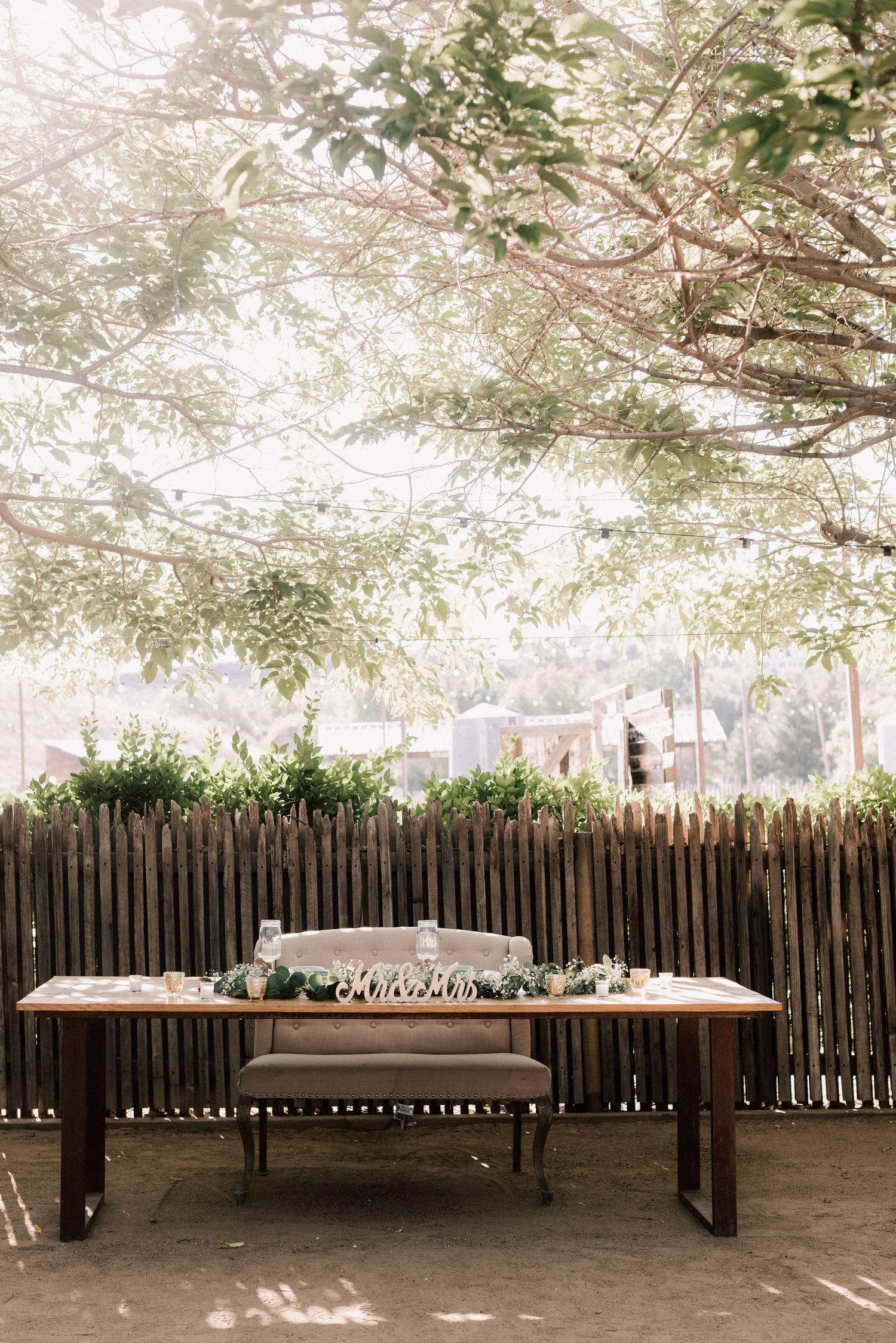 Rustic outdoor sweetheart table with wooden bench, greenery centerpiece, and candles beneath a lush tree canopy.