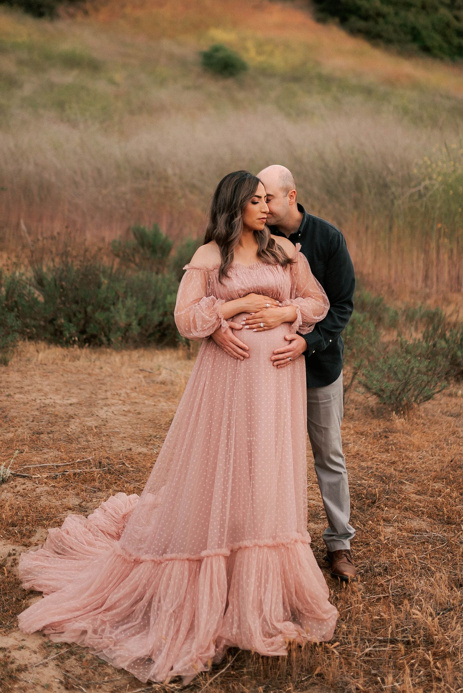 Couple sharing a tender maternity photo in a field, woman in pink tulle gown cradling baby bump.