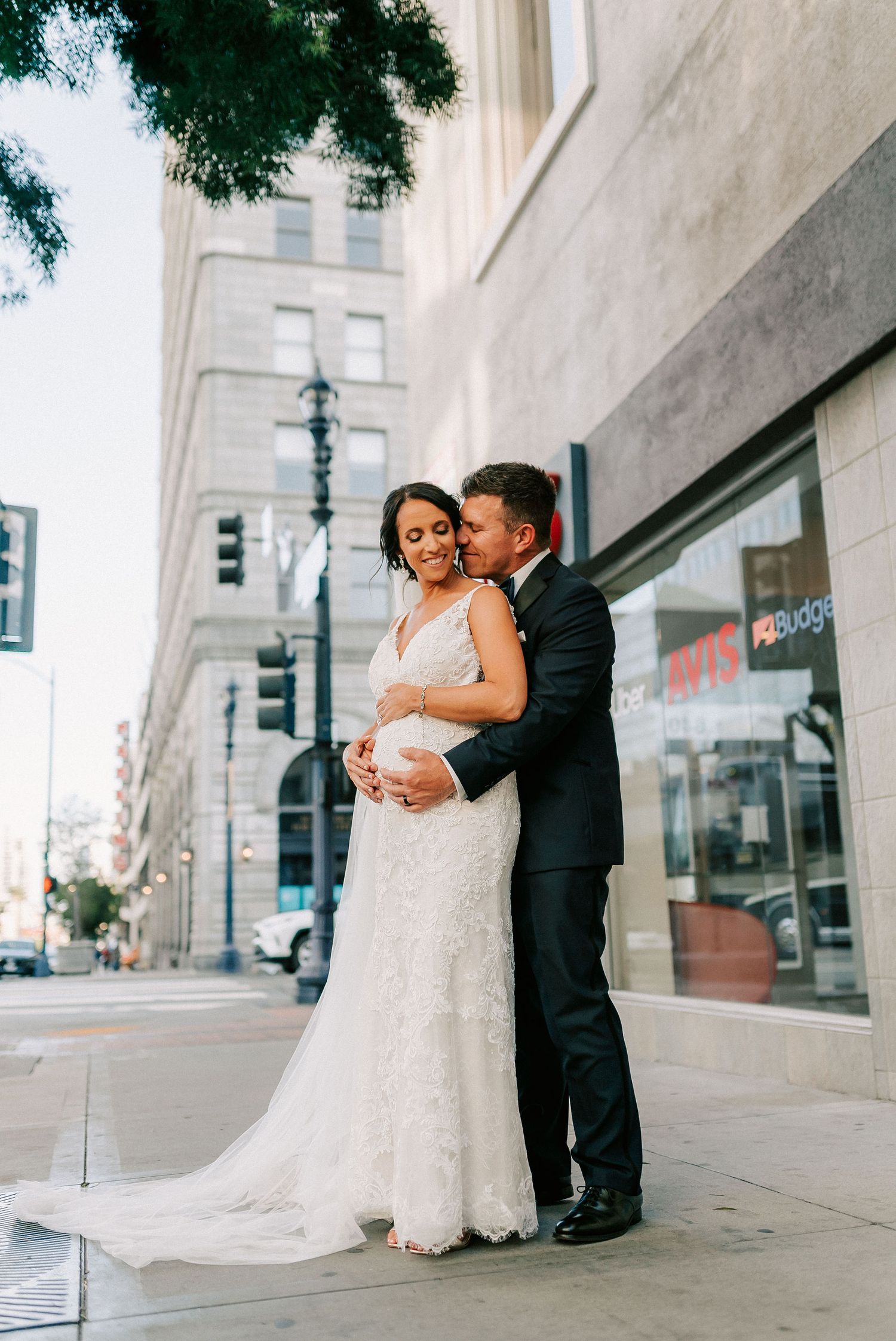 Bride and groom embrace on a city sidewalk, she in a lace gown, he in a dark suit, sharing a tender moment.