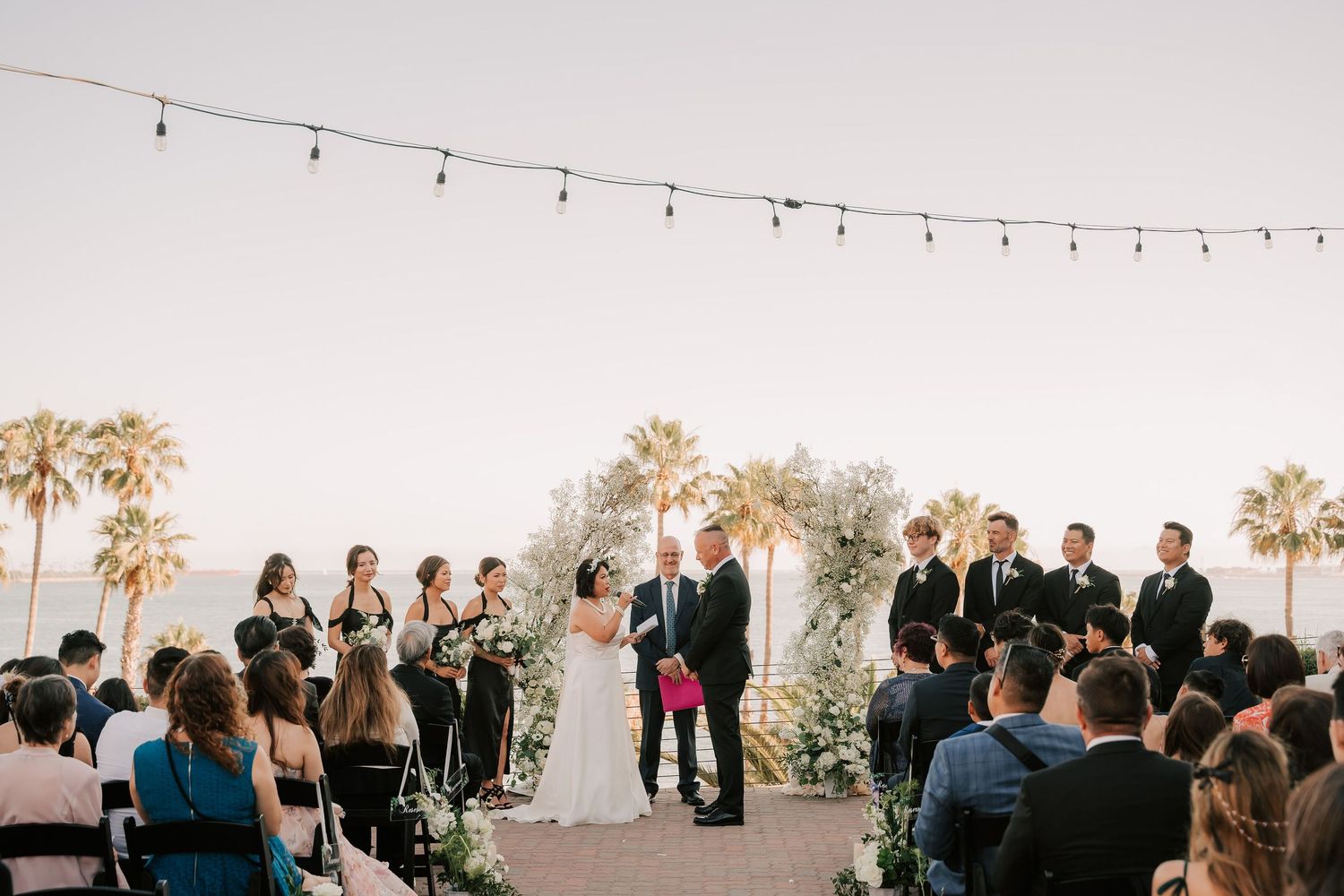 Outdoor beach wedding ceremony with bride and groom at floral arch, bridesmaids, groomsmen, and guests under string lights at sunset.