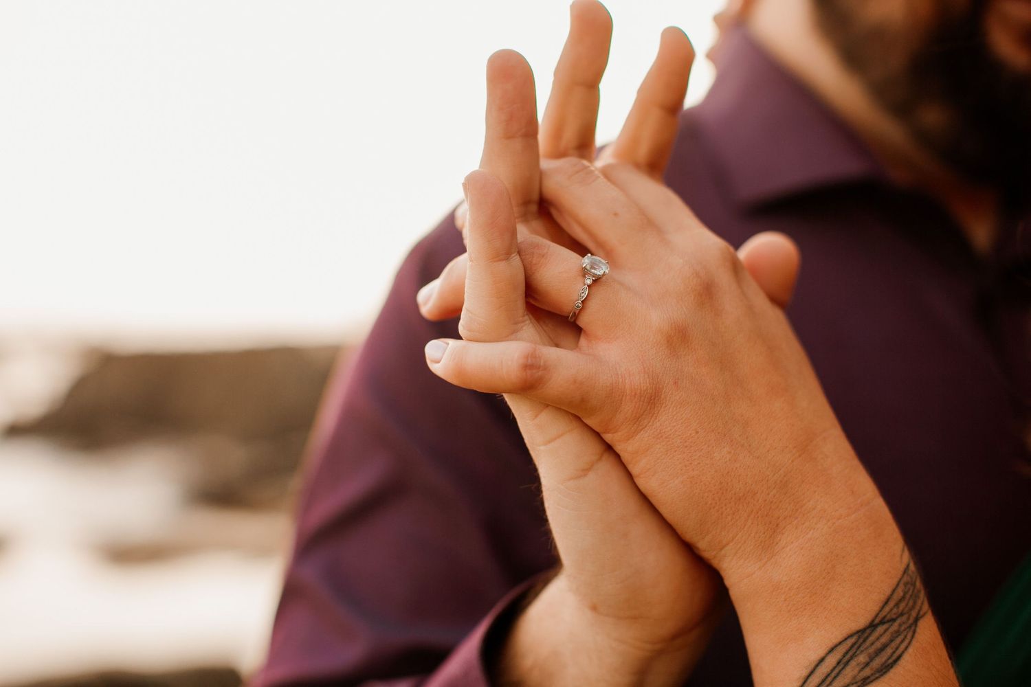 Close-up of couple holding hands showing diamond engagement ring, with tattoo visible on wrist outdoors.