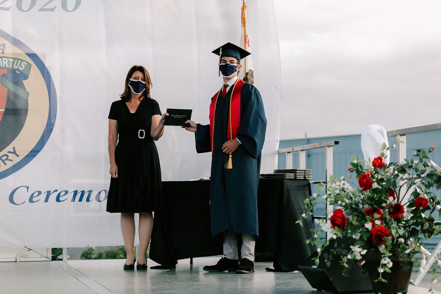 Graduate in cap and gown receives diploma at outdoor graduation ceremony while both wear protective face masks.