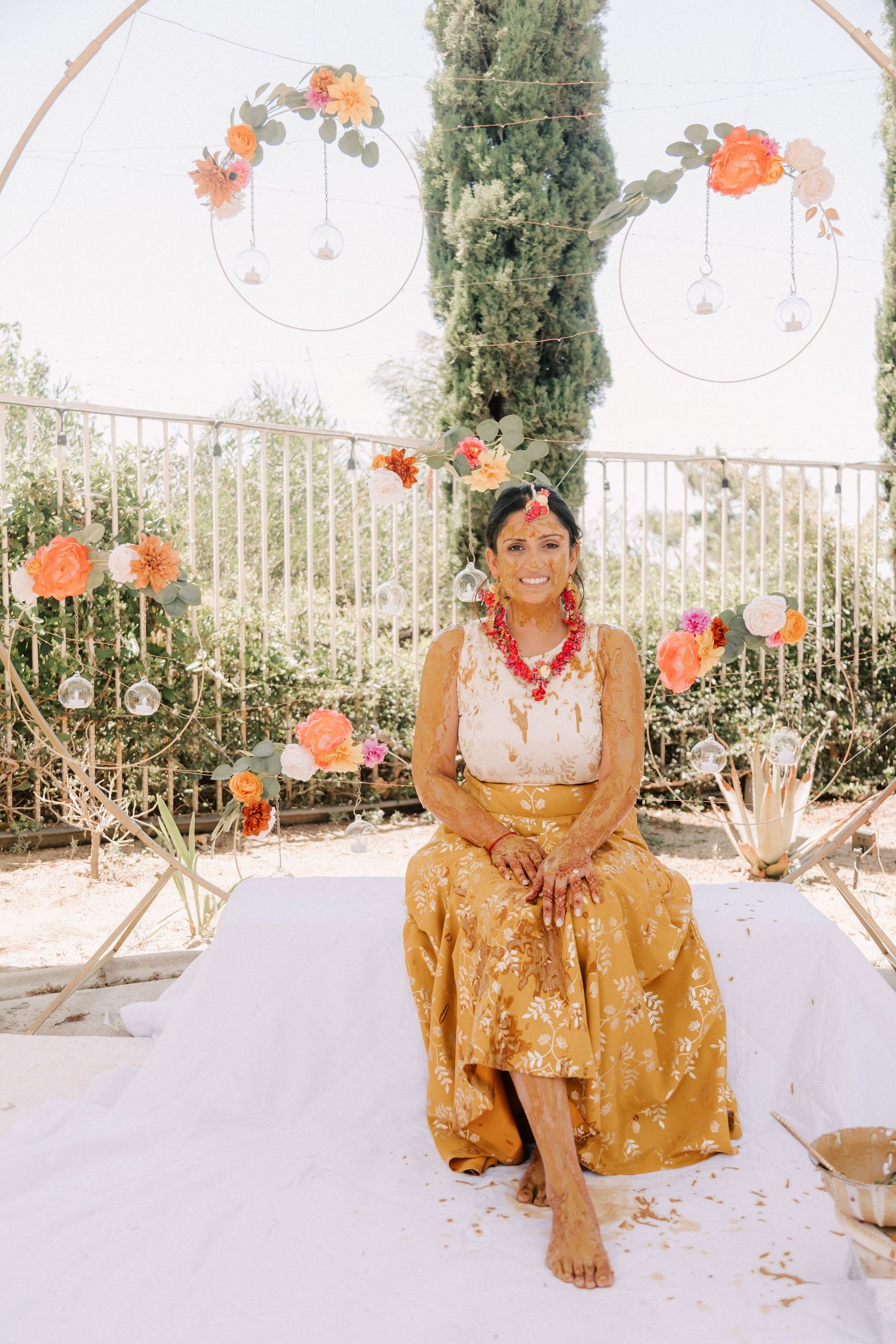 Bride in gold skirt and white top sits barefoot before floral arch at Indian haldi ceremony.
