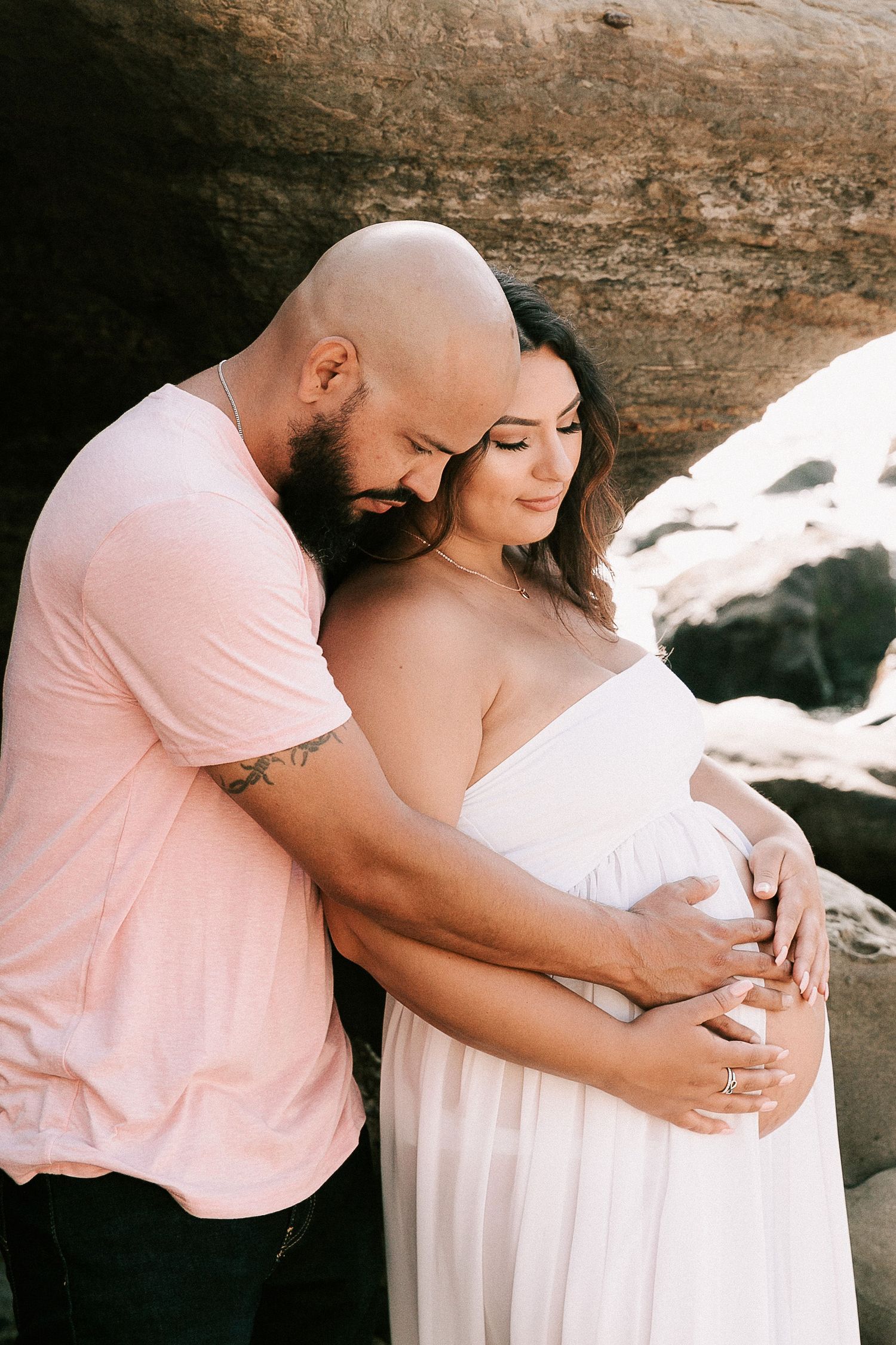 Man in pink shirt tenderly embracing pregnant woman in white dress against rocky background.